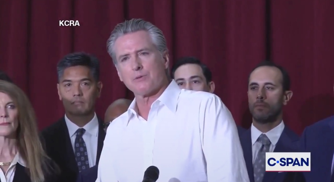 California Governor Gavin Newsom speaks at a press conference surrounded by officials, addressing key issues with a serious expression and a microphone in hand.