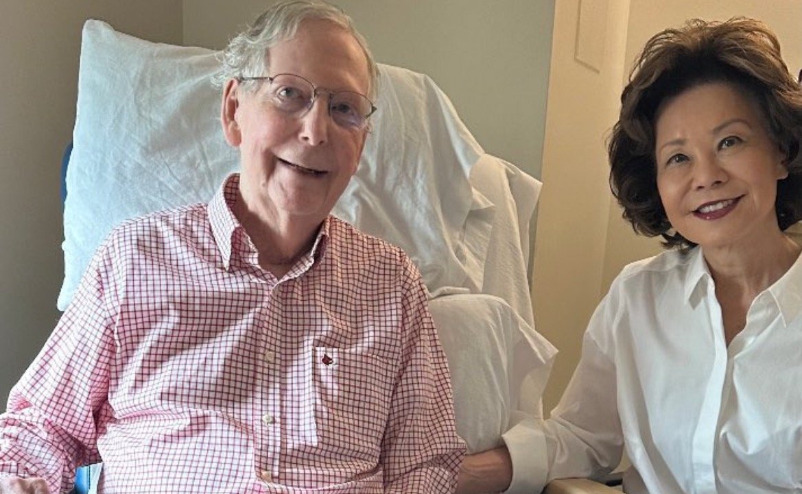 A smiling elderly man in a hospital setting poses for a photo with a woman, both appearing friendly and engaged.