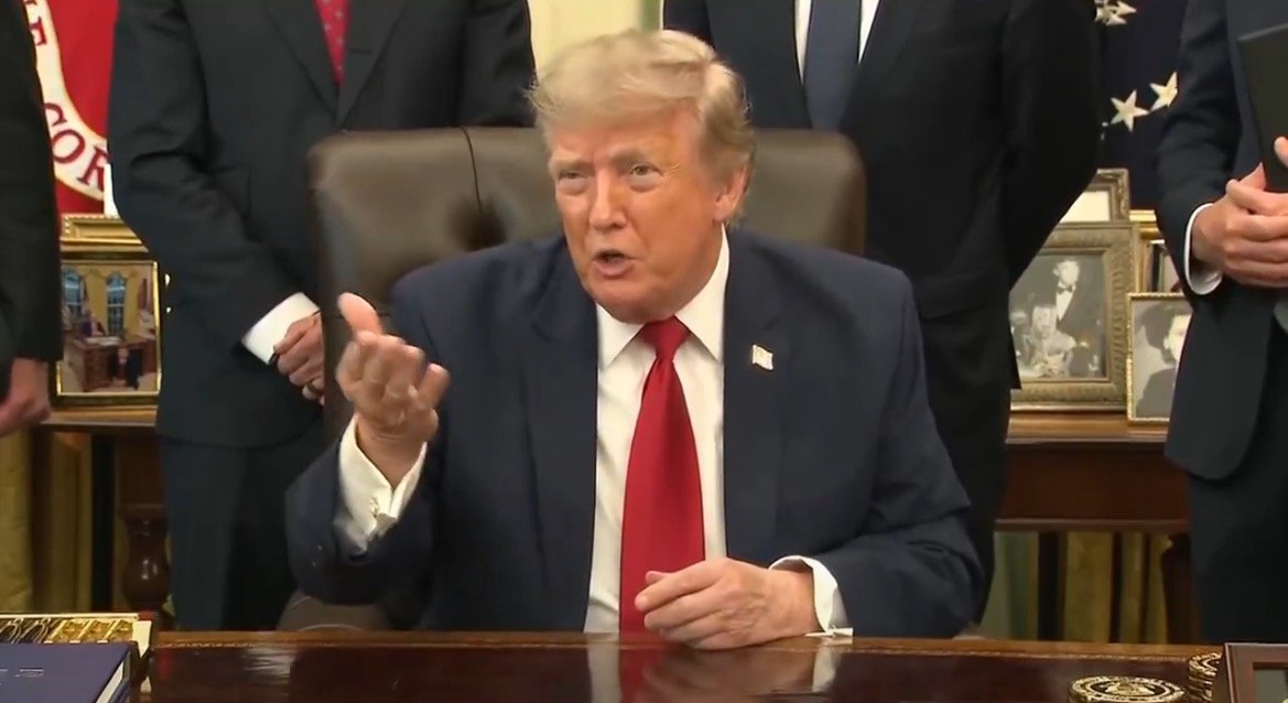 Donald Trump gestures while speaking in the Oval Office, surrounded by officials and framed photographs, during a formal event.
