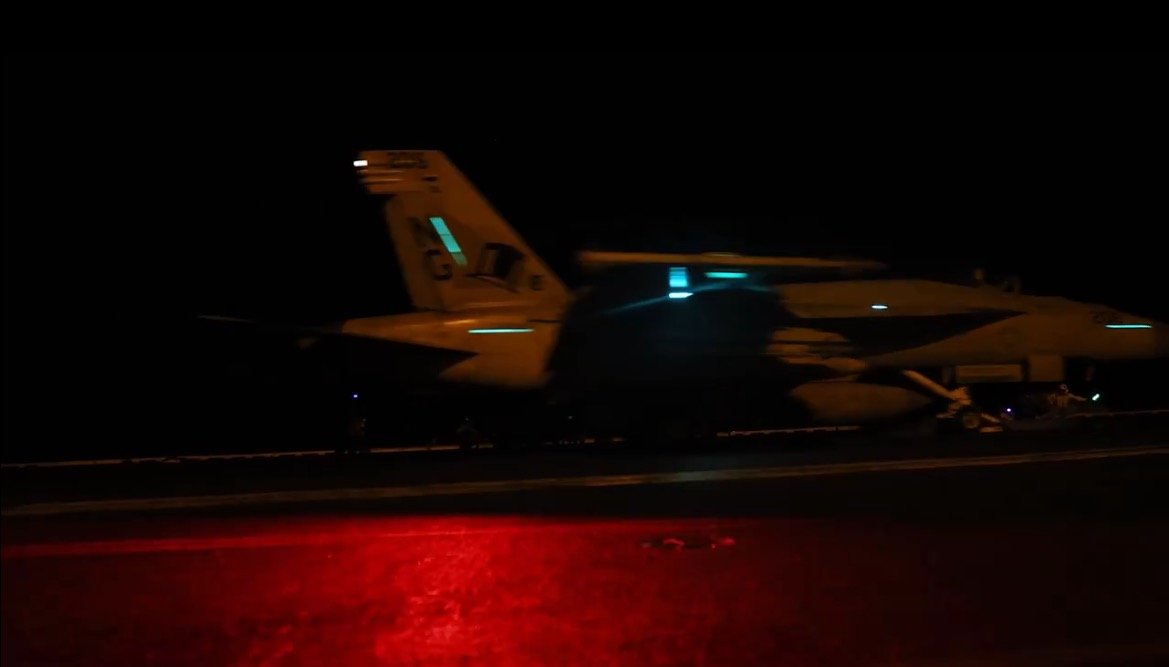 Fighter jet launching from an aircraft carrier deck at night, illuminated by red and blue lights.