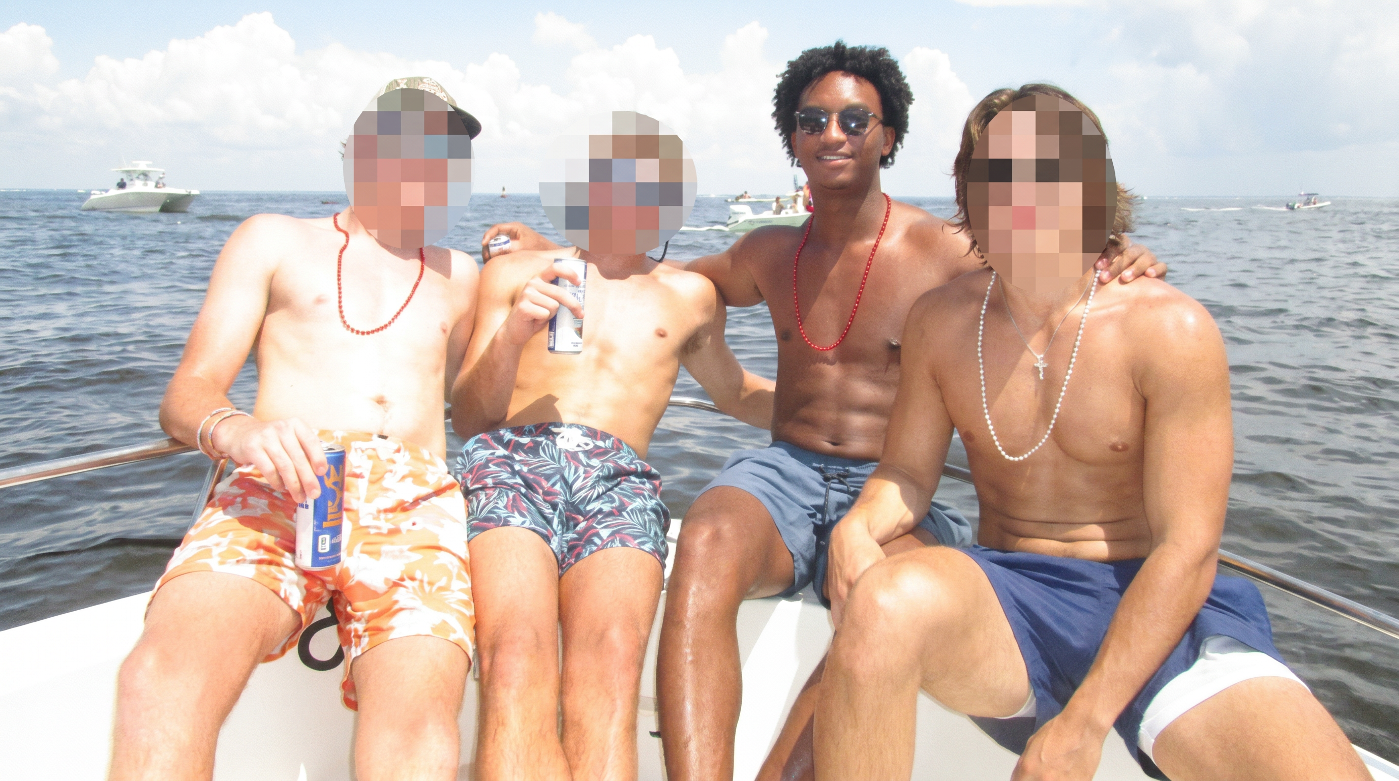 Four shirtless young men in swim trunks sitting on the edge of a boat on open water, faces pixelated.
