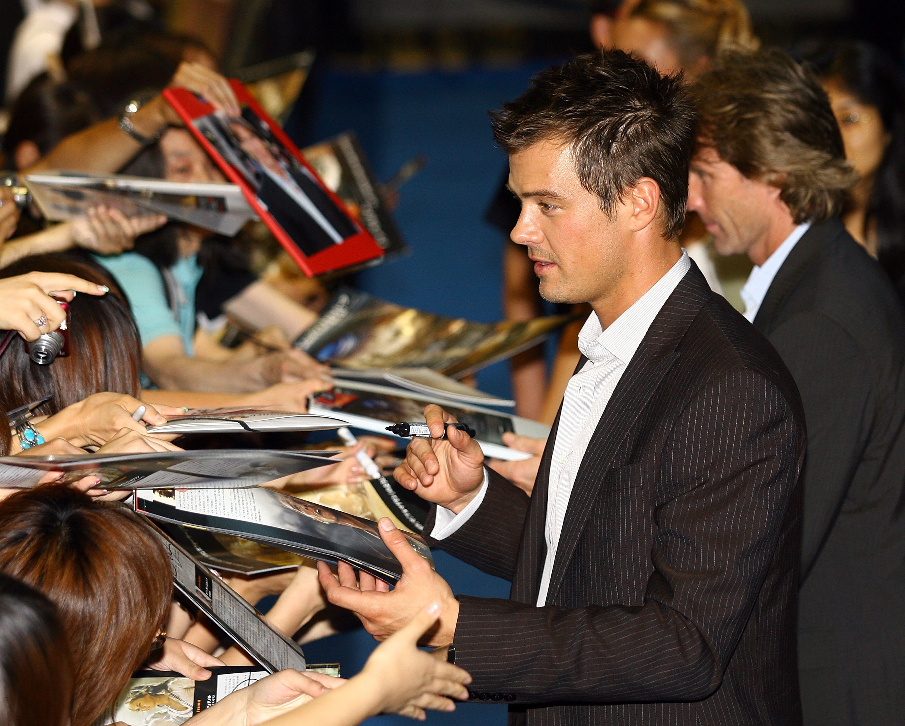 A man in a dark pinstripe blazer signing autographs for a large crowd of fans at a movie premiere in Tokyo.