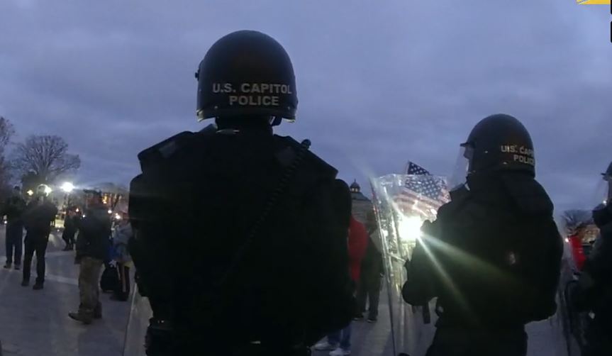 U.S. Capitol Police officers stand guard during a protest at the Capitol, with a cloudy sky and a crowd visible in the background.