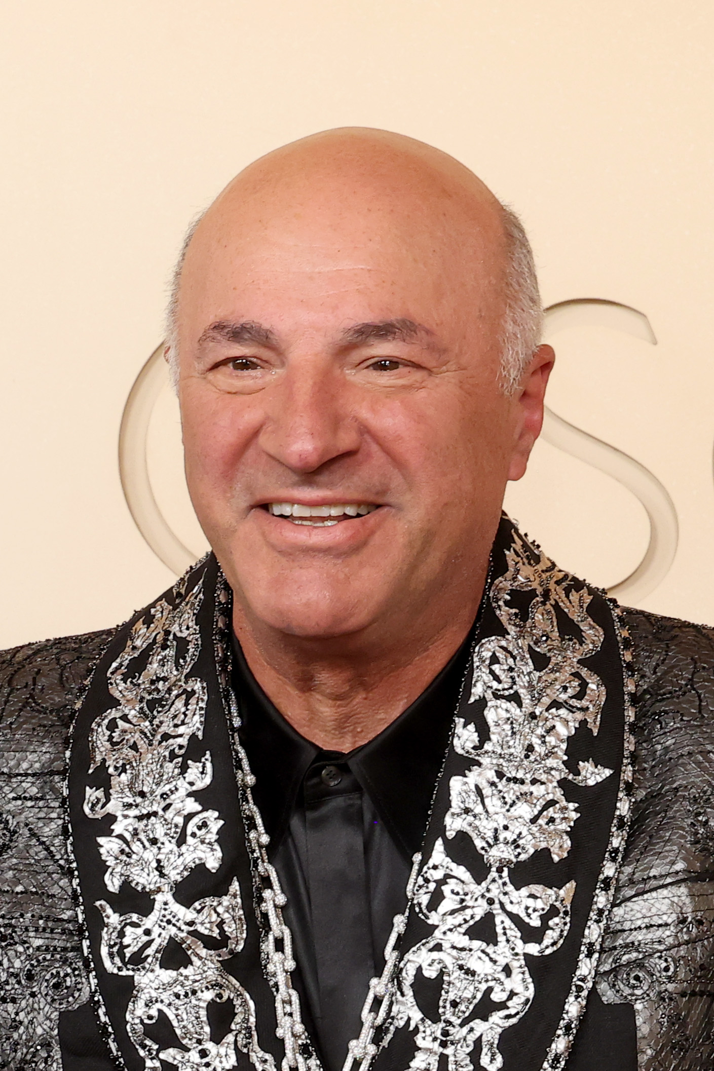 Bald older man in a black shirt and silver-embroidered black blazer smiling at a red carpet event with a decorative logo backdrop.