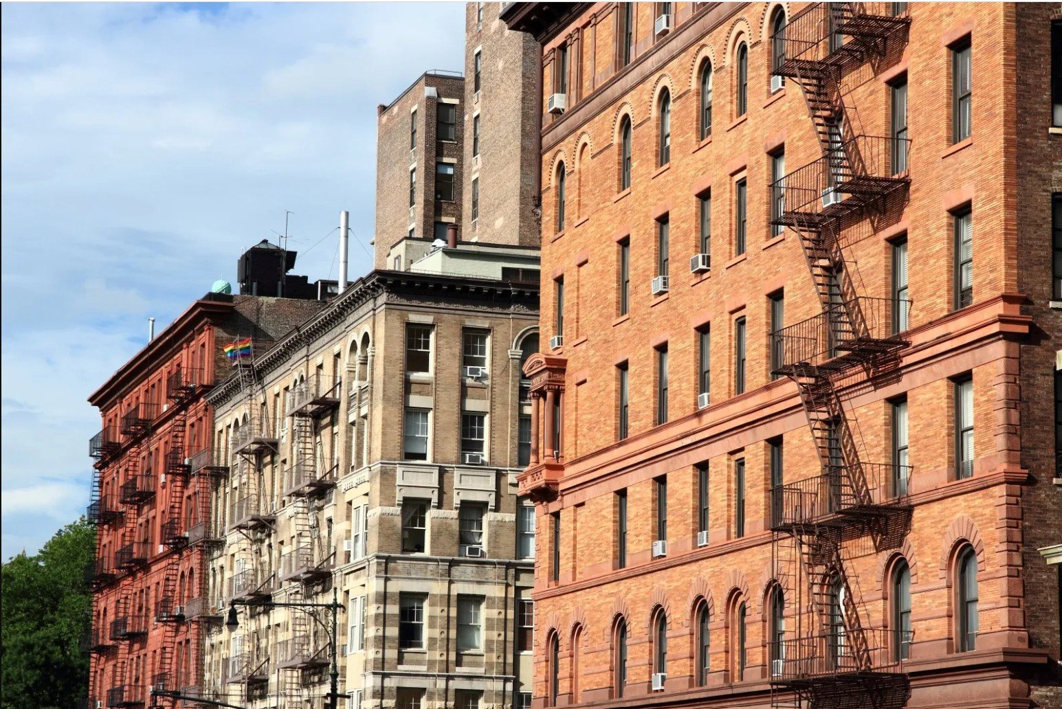 Historic buildings with fire escapes and diverse architectural styles in an urban setting under a partly cloudy sky.