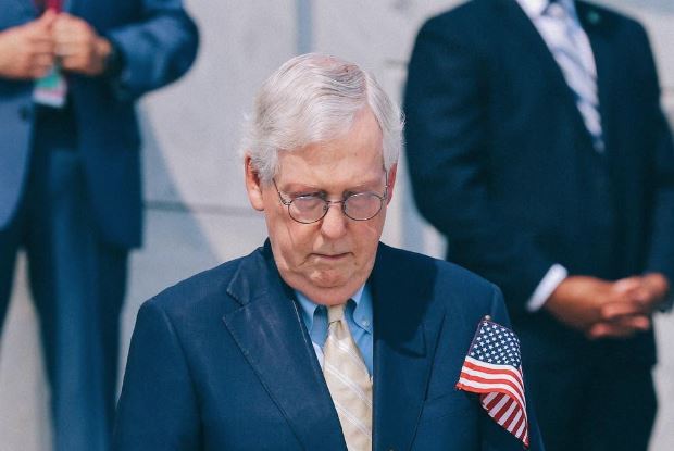 Mitch McConnell, wearing a suit and glasses, stands with his head bowed and an American flag pinned to his lapel during a formal event.