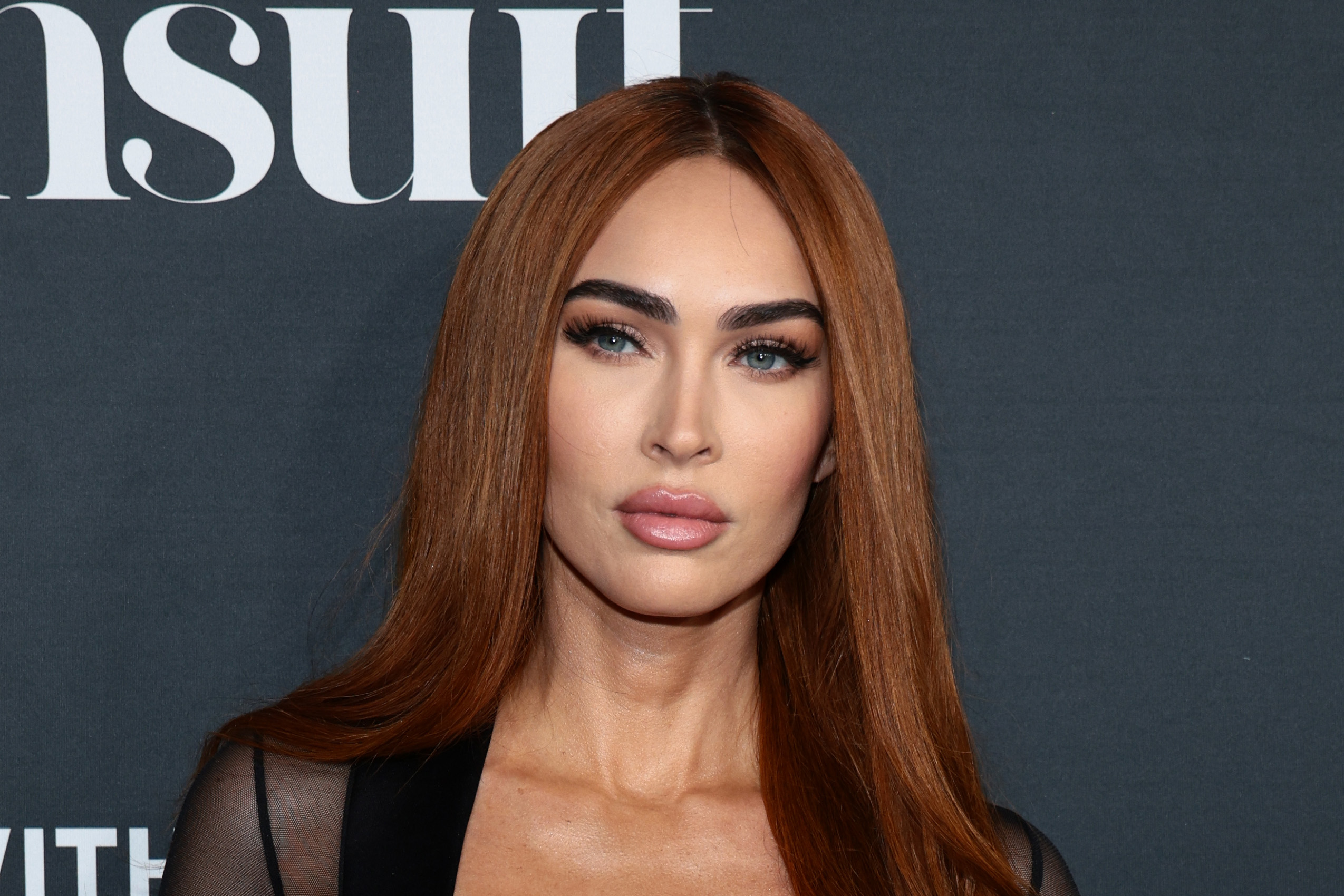 Close-up of a woman with long straight auburn hair, dramatic brows, blue eyes, and nude makeup in front of a gray swimsuit event backdrop.