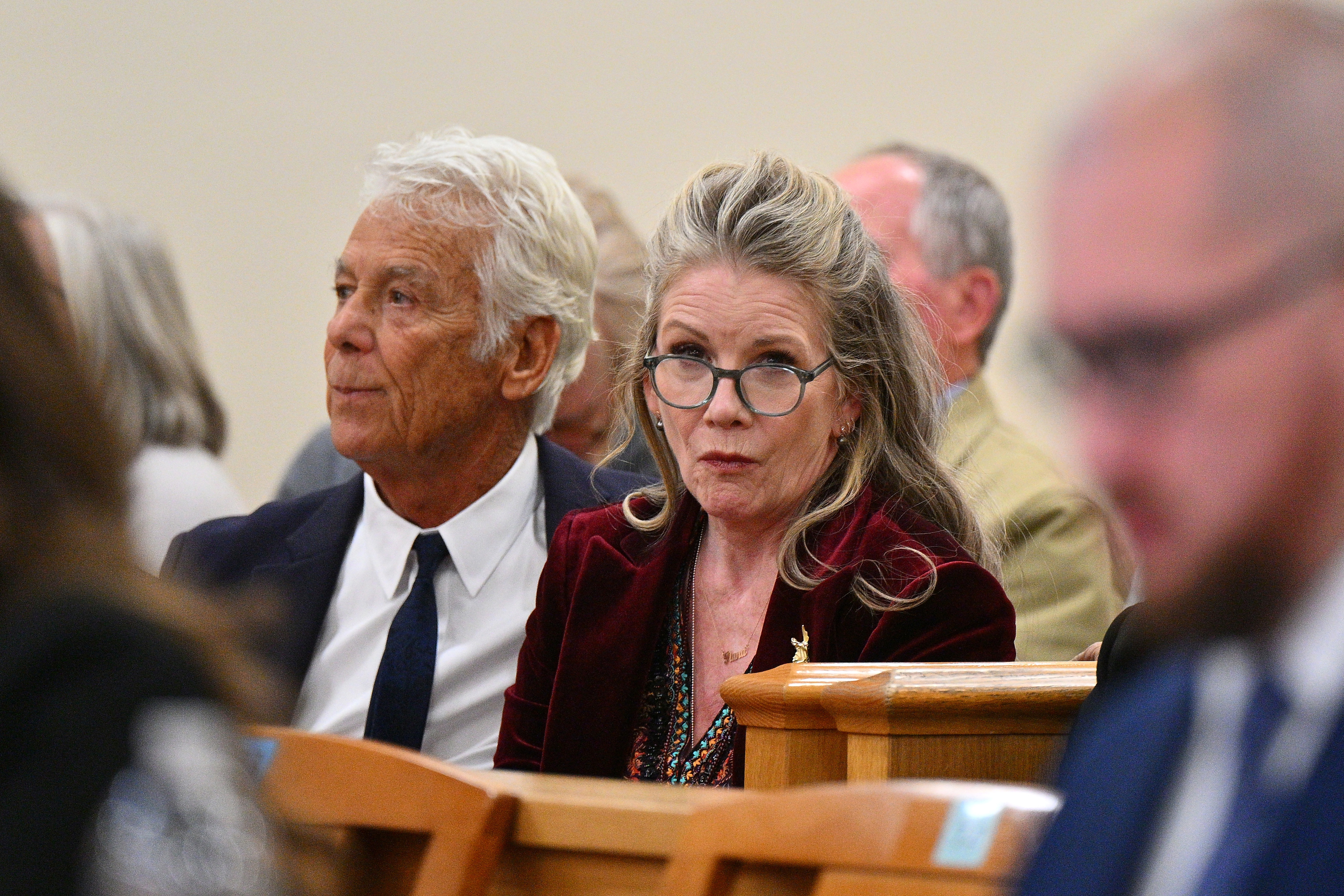 Melissa Gilbert wearing glasses and a burgundy velvet jacket sits beside an older man in a navy suit inside a courtroom.