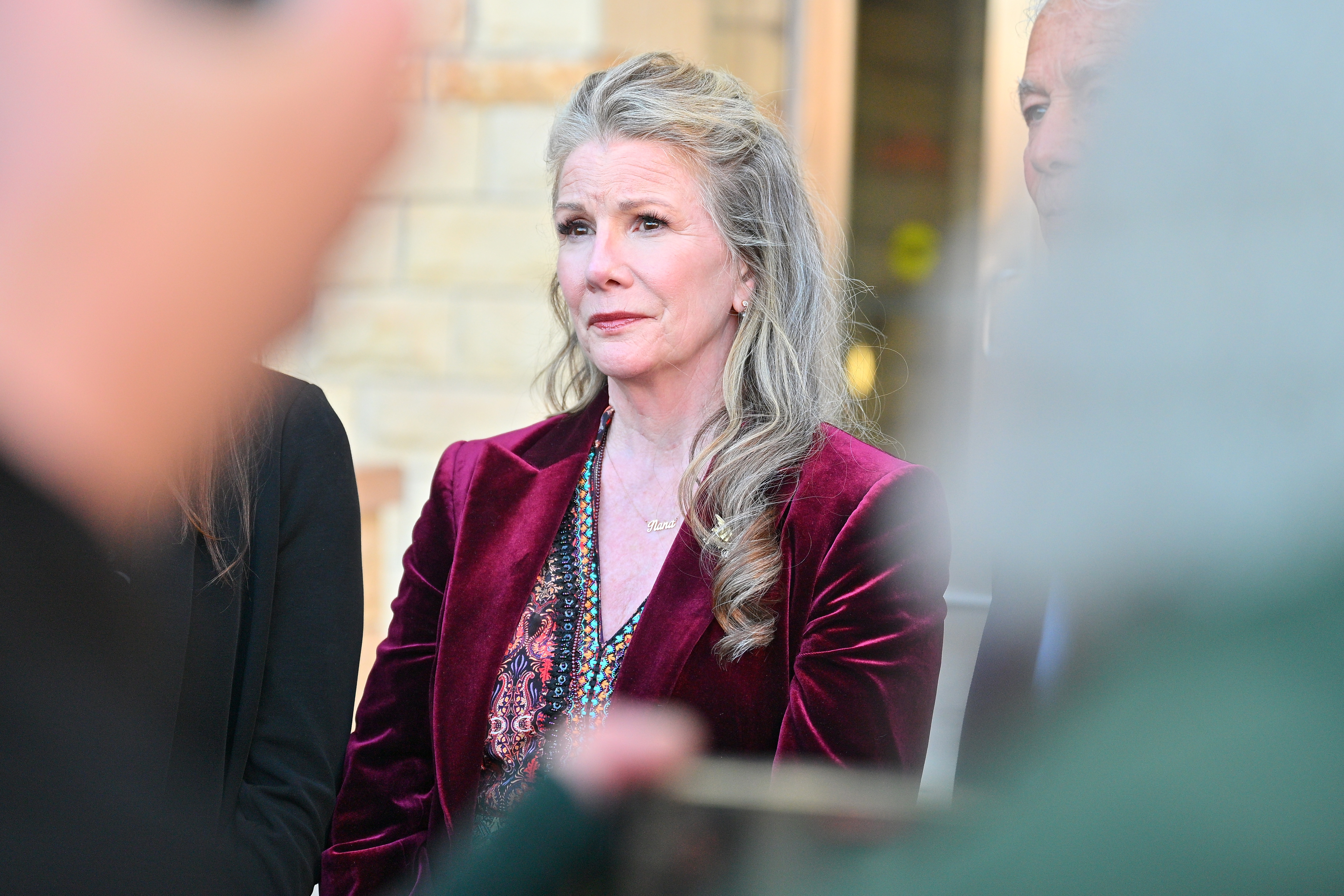 A woman with long gray-blonde hair in a burgundy velvet blazer stands outdoors, looking ahead with a somber expression.