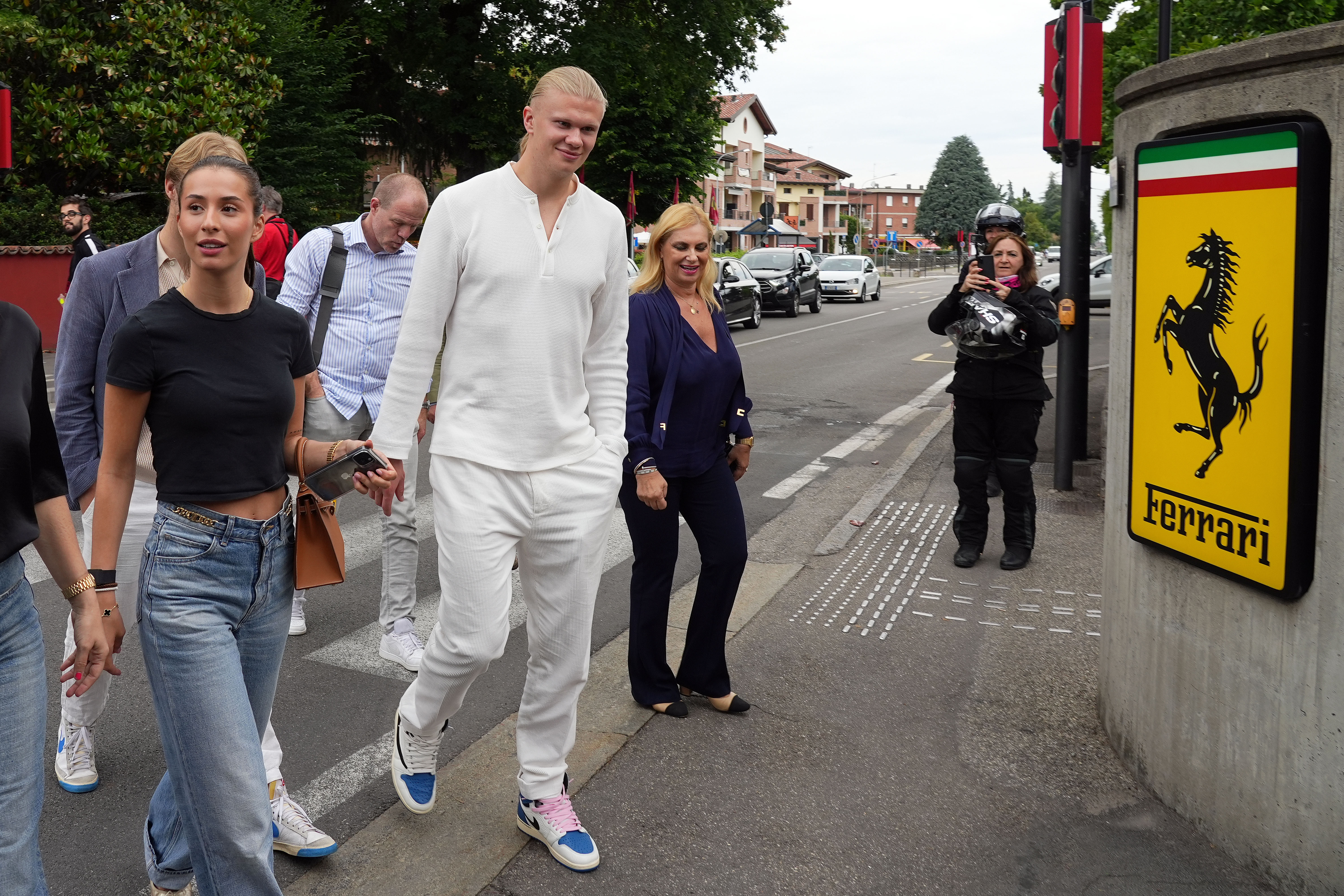 Erling Haaland in an all-white outfit and Isabel Haugseng Johansen in a black crop top and jeans walk past a yellow Ferrari logo sign in Maranello, Italy.
