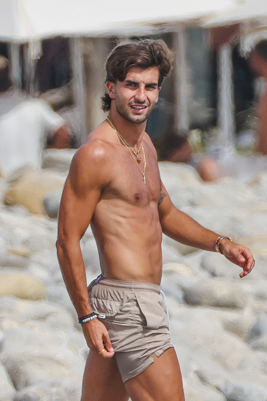 Shirtless young man with dark curly hair and gold necklaces smiling on a rocky beach.