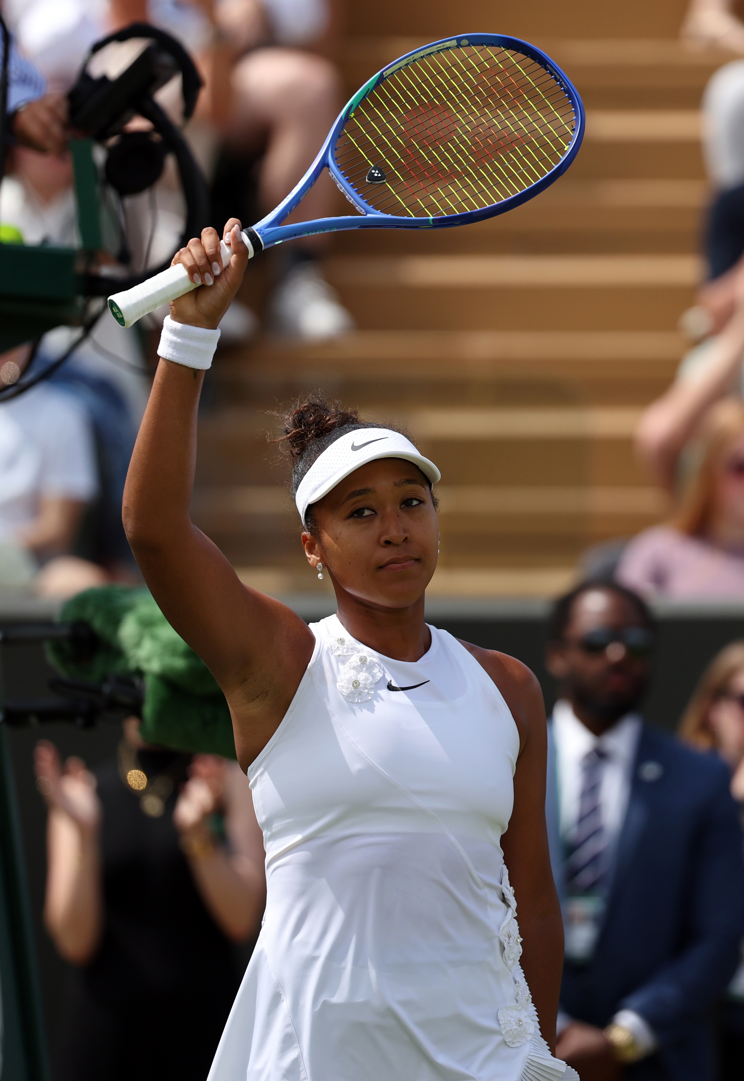 Naomi Osaka in a white Nike dress and visor raises her blue Wilson racket overhead toward the crowd at Wimbledon.