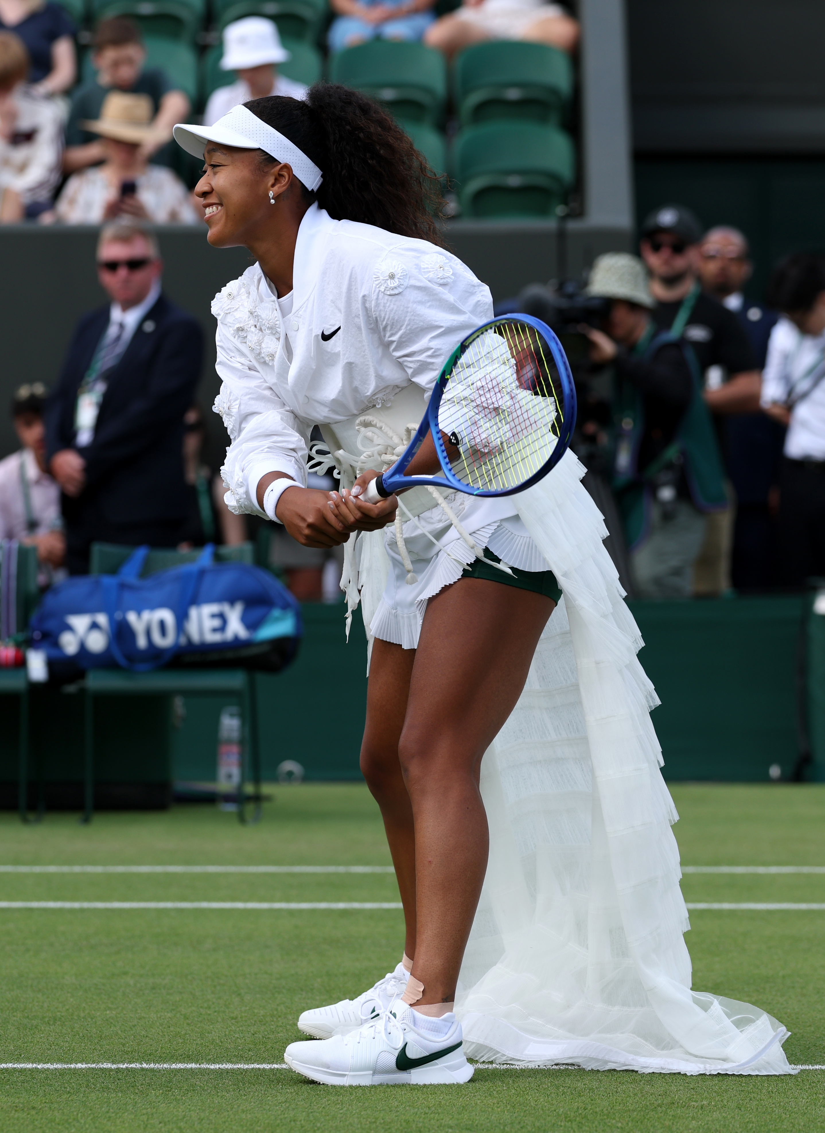 Naomi Osaka smiling and bent forward on a grass court in a white floral ruffled outfit, holding a blue tennis racket.