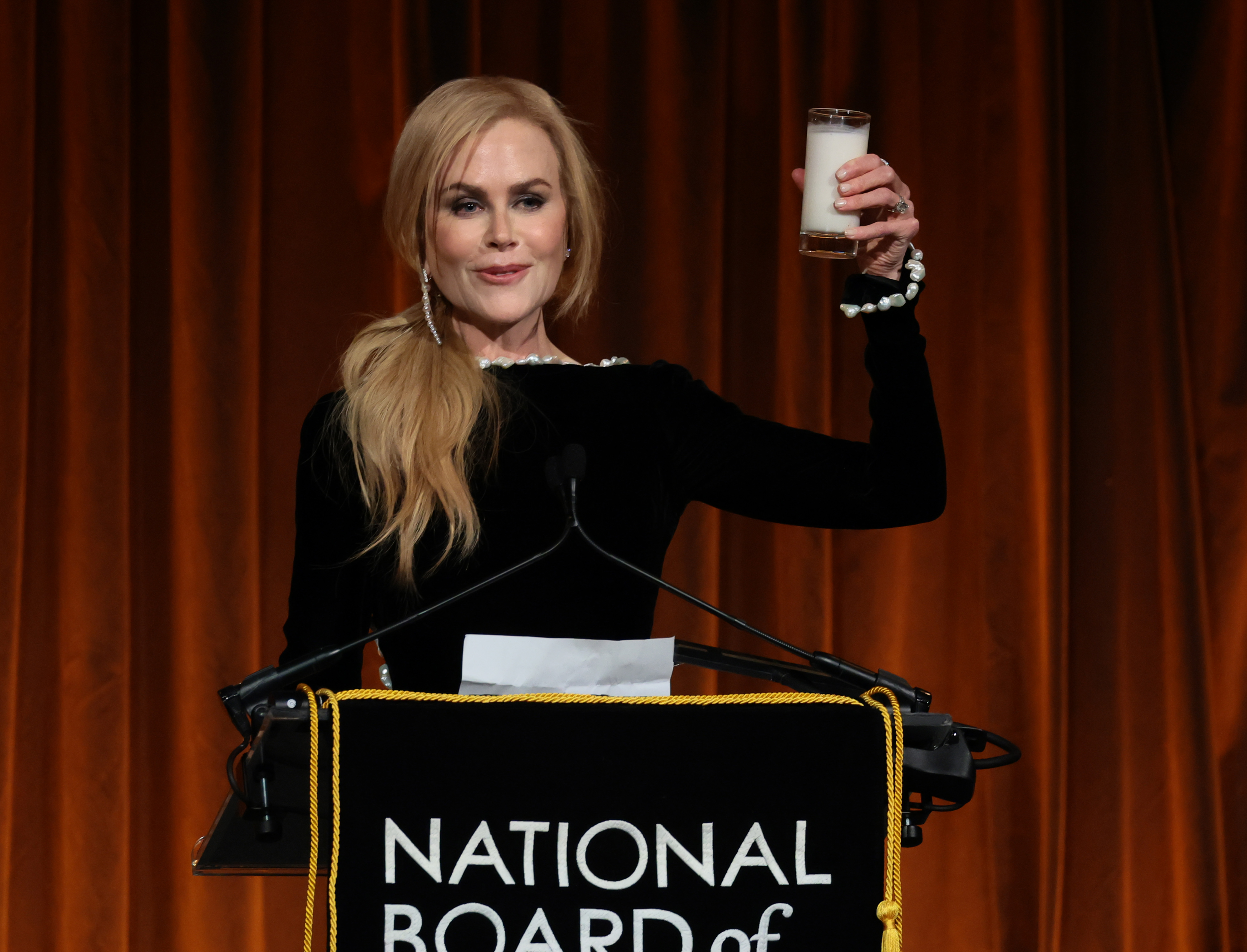 Nicole Kidman in a black dress holds a glass aloft at a podium reading 'National Board of Review' on stage.