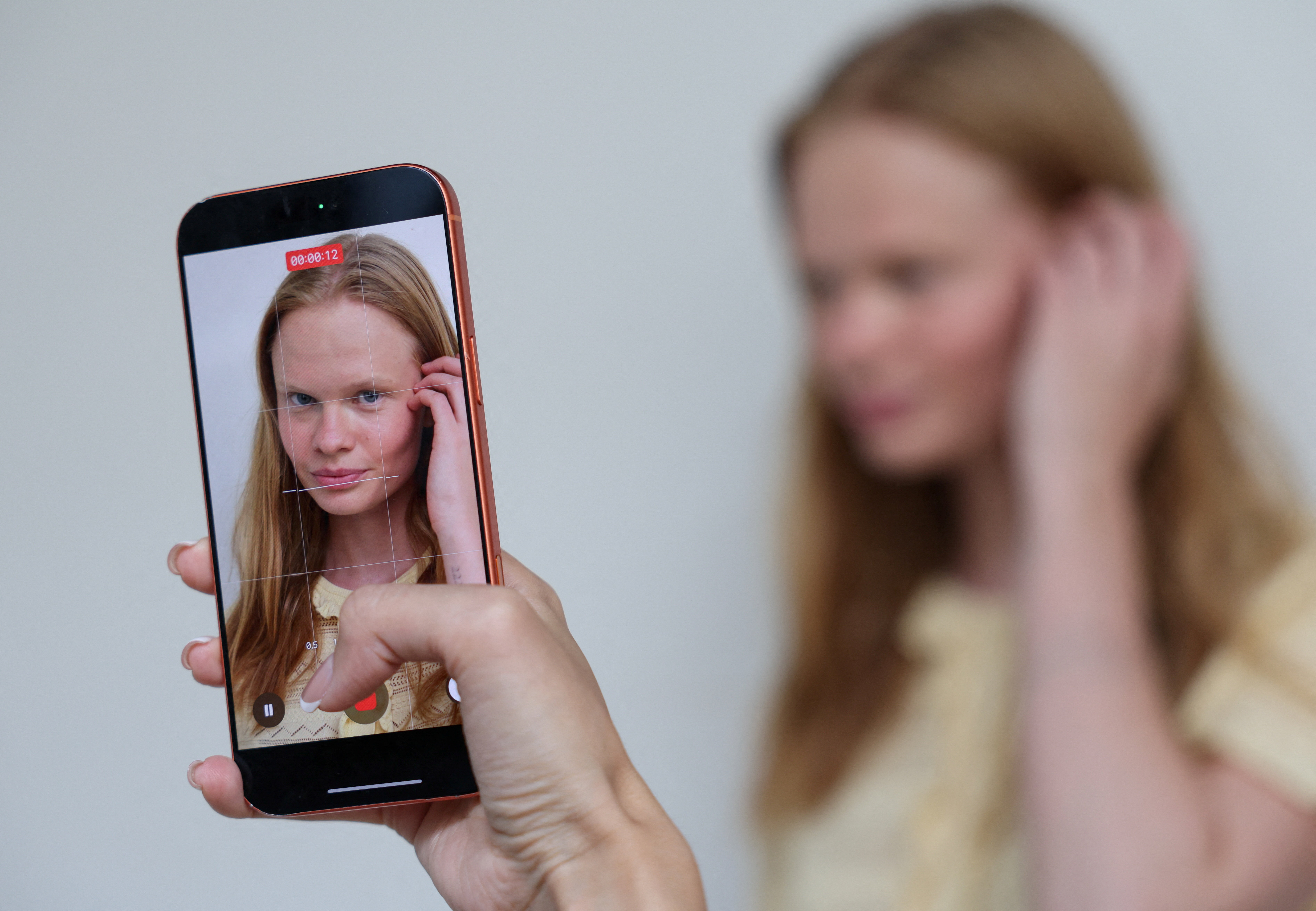 A smartphone held up showing a video recording of a young woman with long blonde hair touching her ear, with the blurred subject visible in the background.