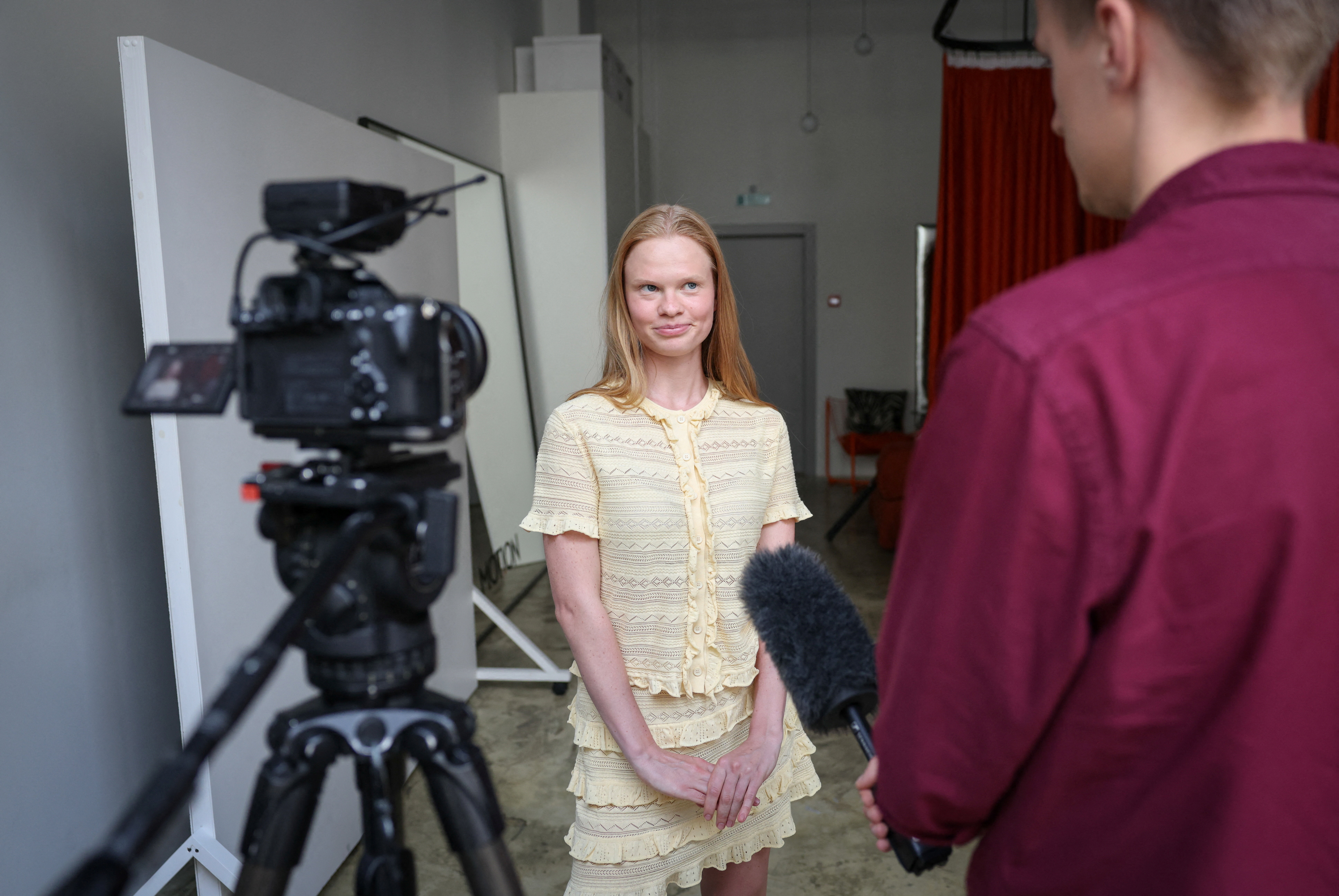 A young blonde woman in a yellow lace outfit stands in a studio being interviewed, with a video camera on a tripod to her left and a person holding a microphone to her right.