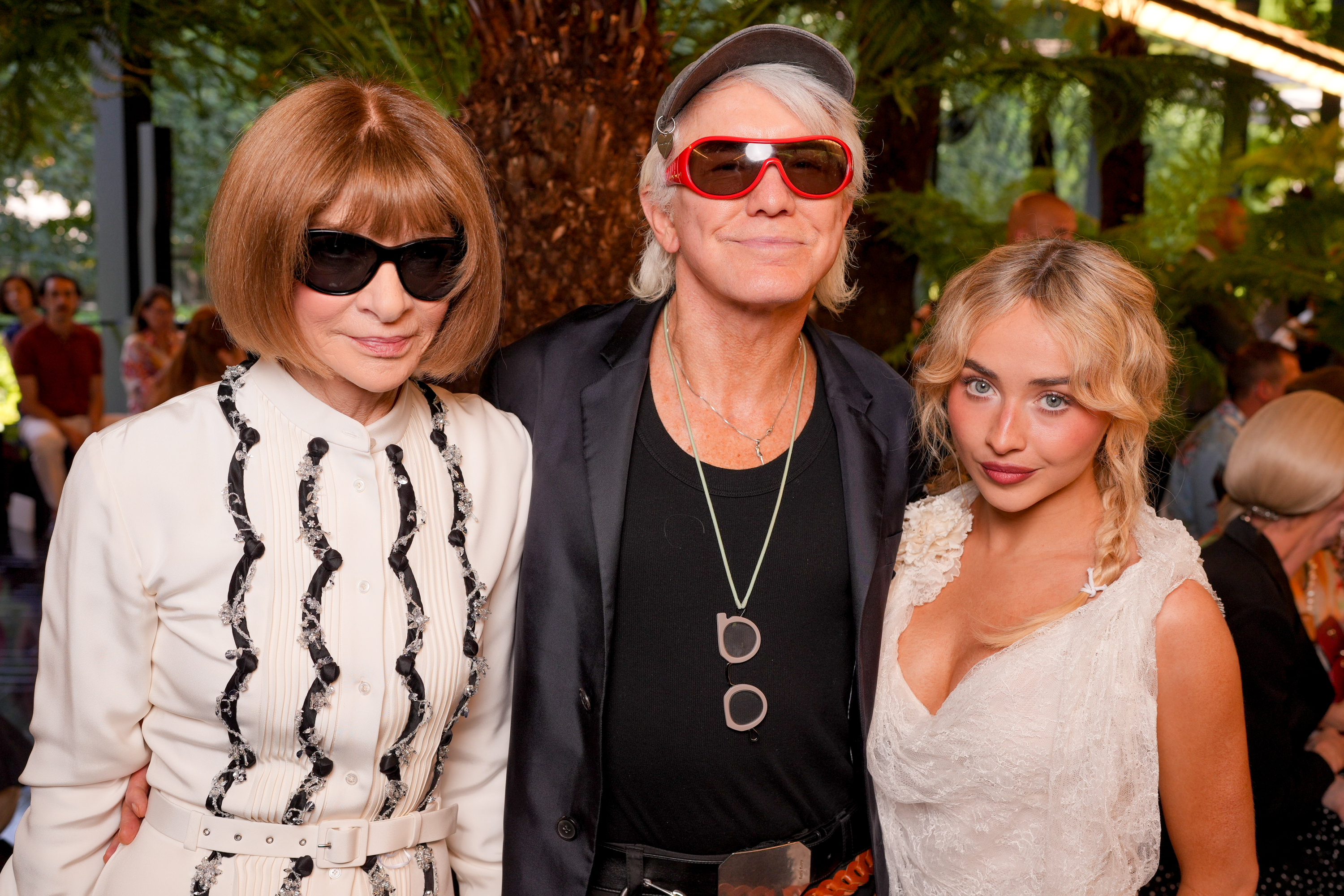 Anna Wintour in black sunglasses and white embellished dress, Baz Luhrmann in red wraparound shades and black blazer, and Sabrina Carpenter in white lace dress pose together outdoors.