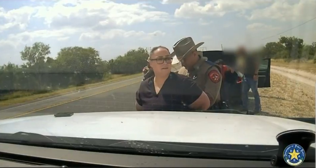 A woman in handcuffs is being apprehended by a Texas state trooper on the roadside, with a clear blue sky in the background.