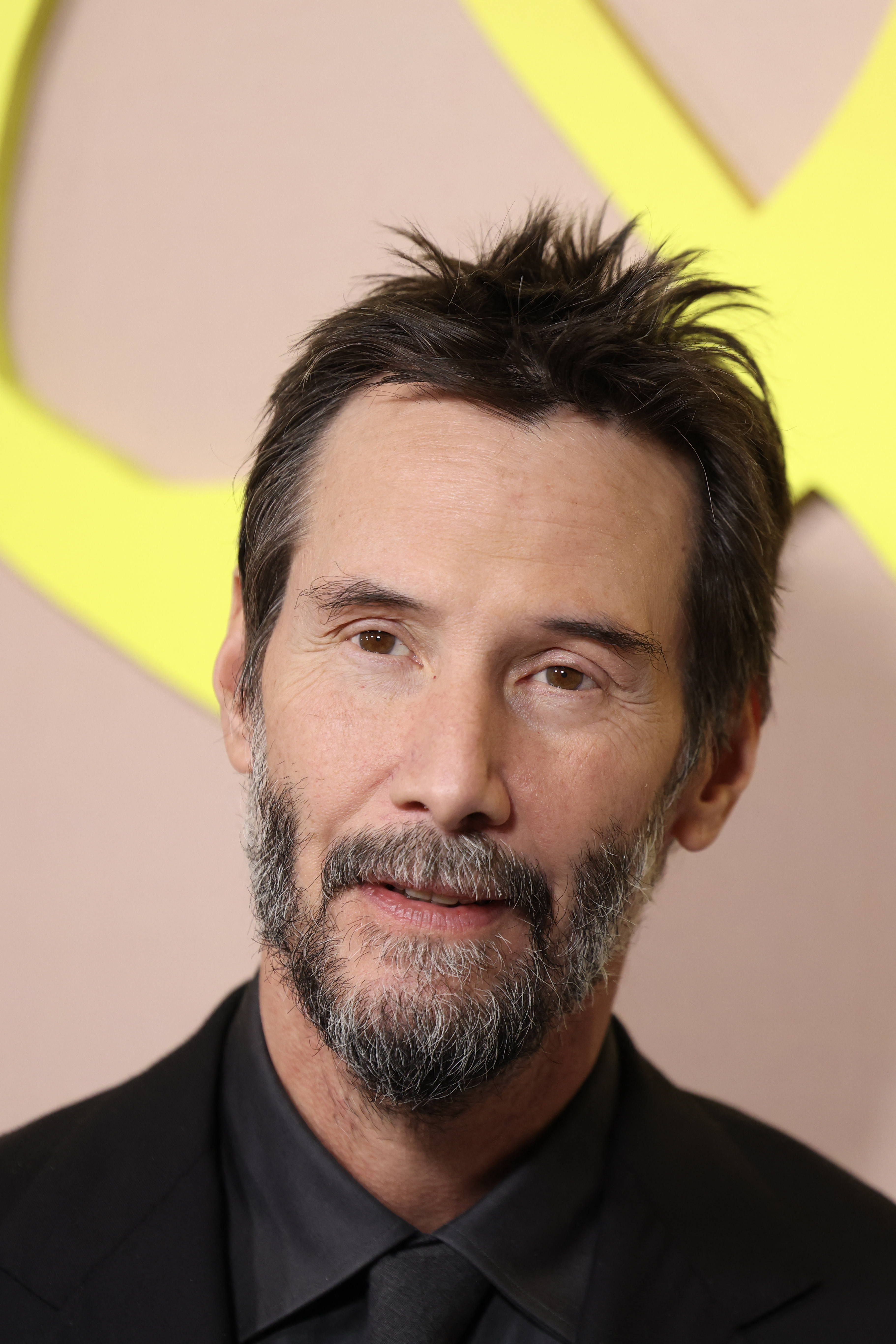 Keanu Reeves with dark spiky hair and a short beard wearing an all-black suit at a premiere event with a yellow logo backdrop.