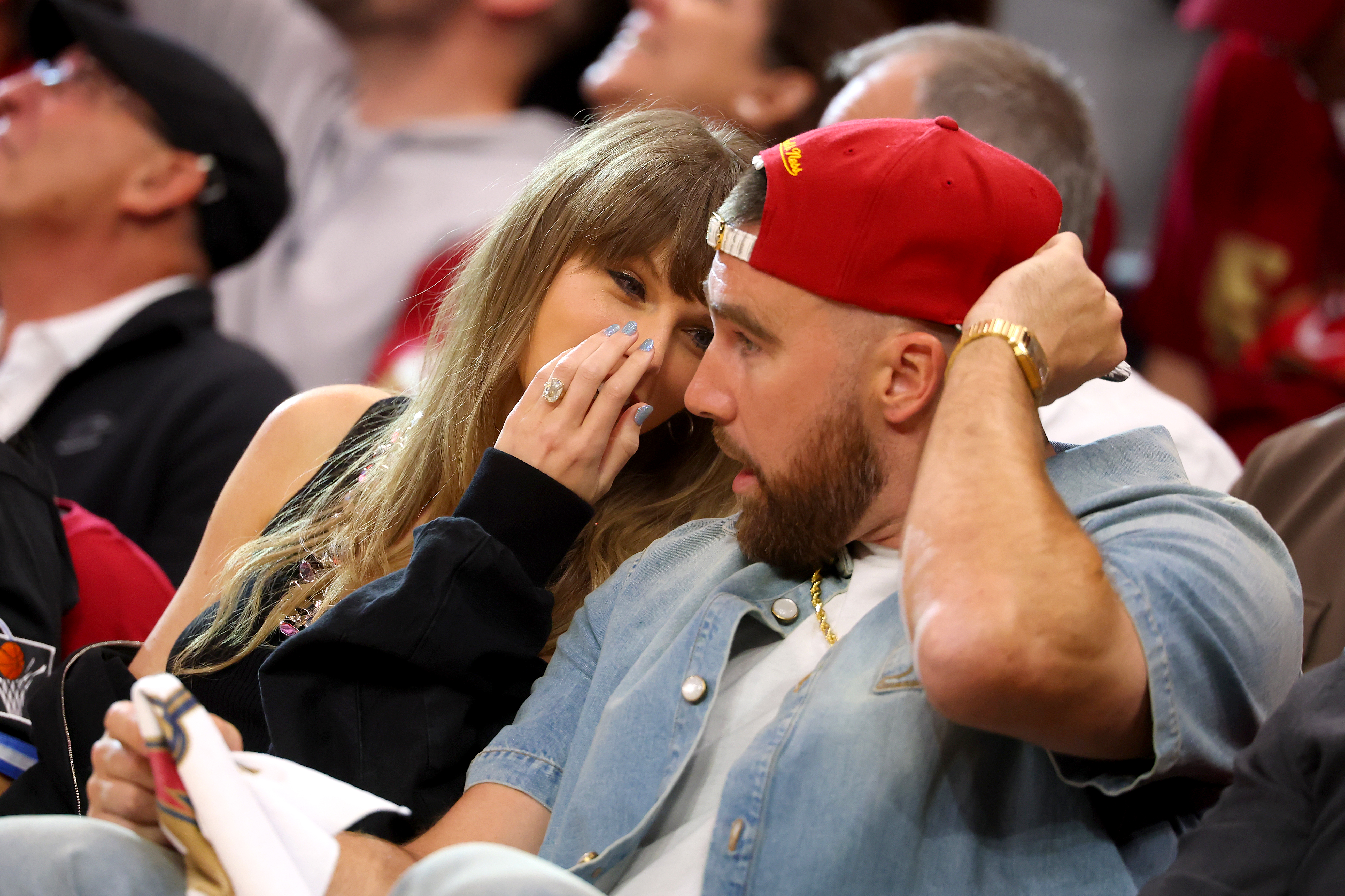 A woman with long wavy hair whispers to a bearded man in a red cap and denim jacket while seated in a crowded basketball arena.