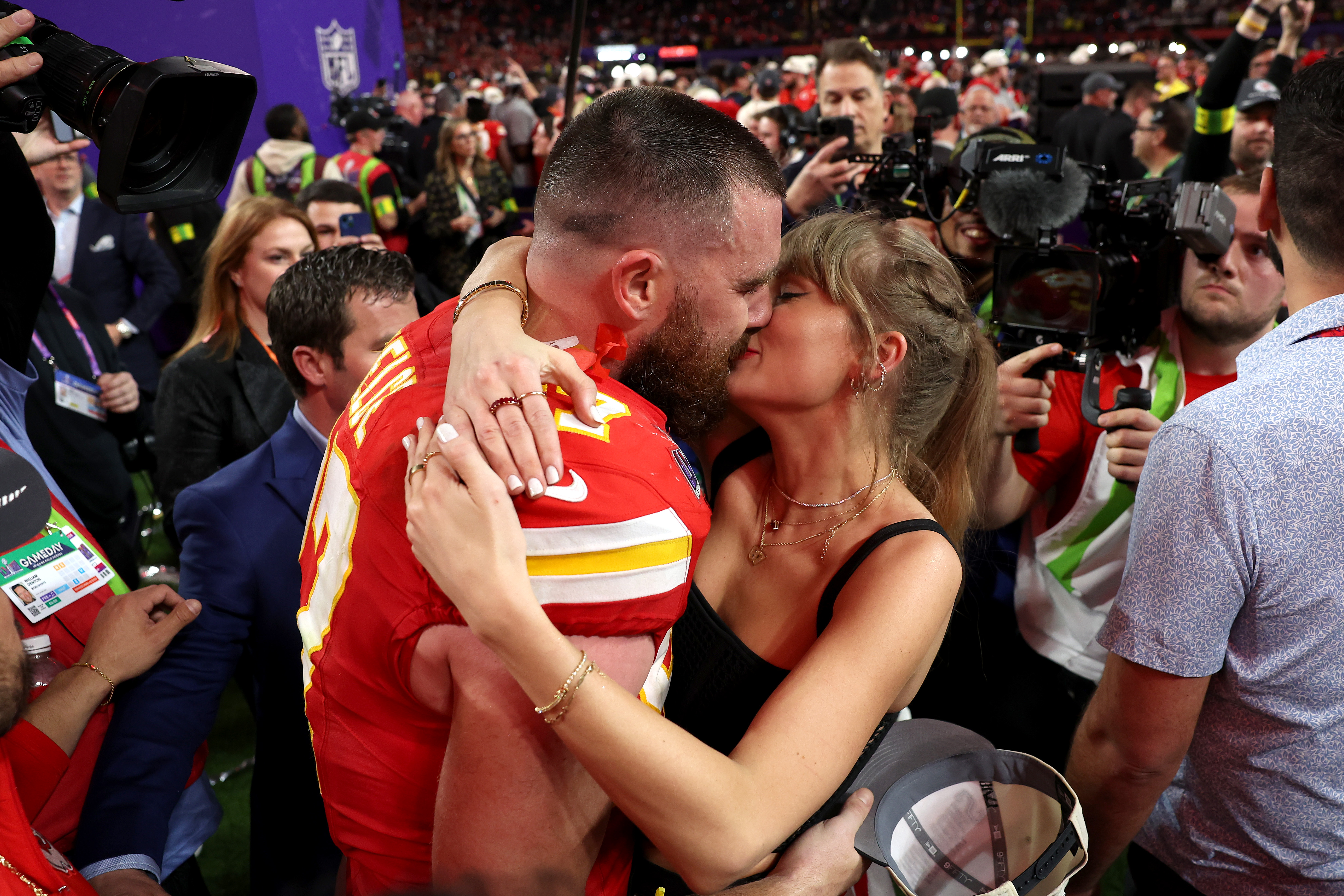 A Kansas City Chiefs player in a red jersey and a woman in a black dress embrace and kiss on a crowded stadium field surrounded by cameras and media.