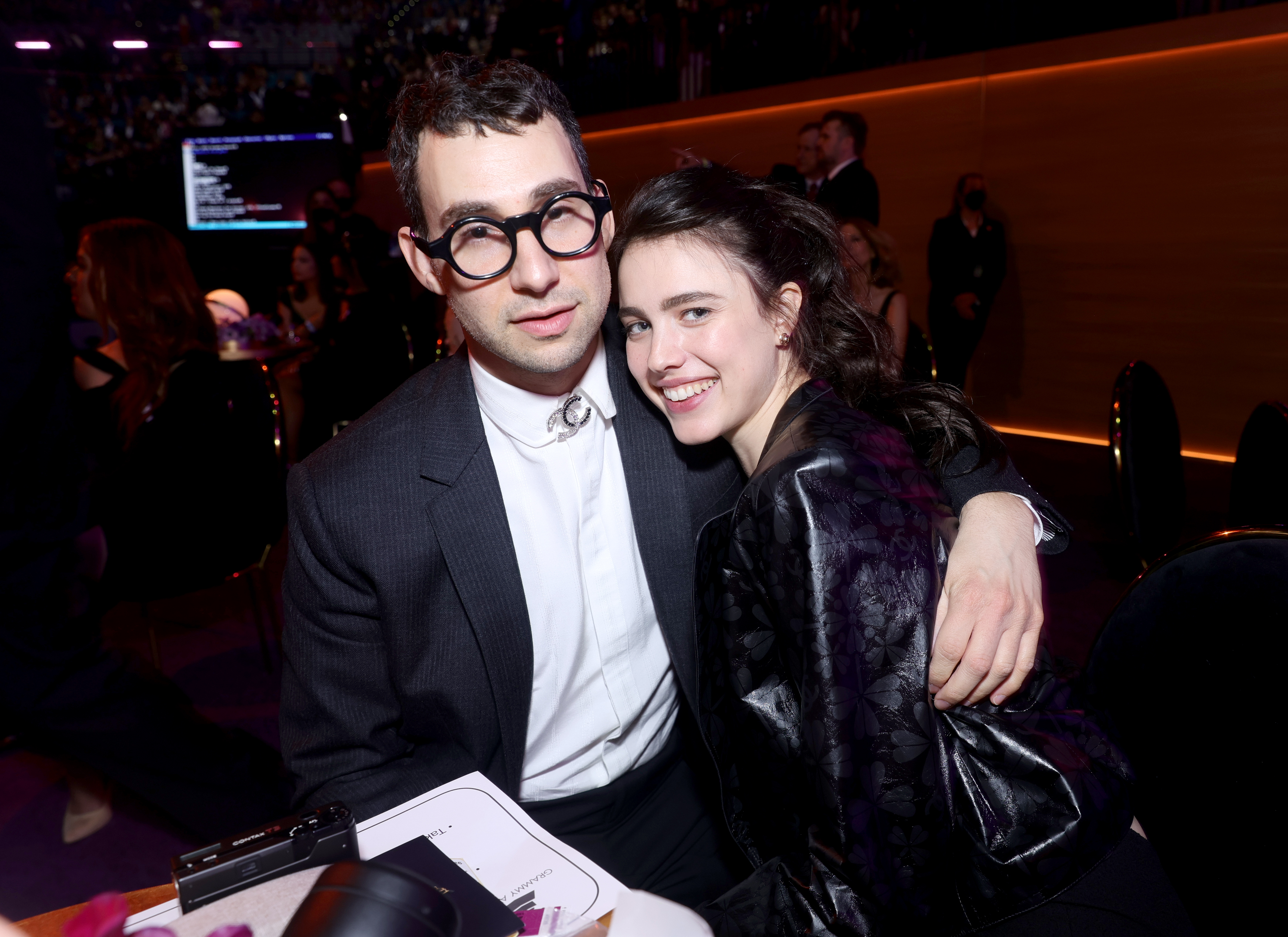 Jack Antonoff in a dark suit with round glasses and Margaret Qualley in a black outfit smile together at the Grammy Awards.