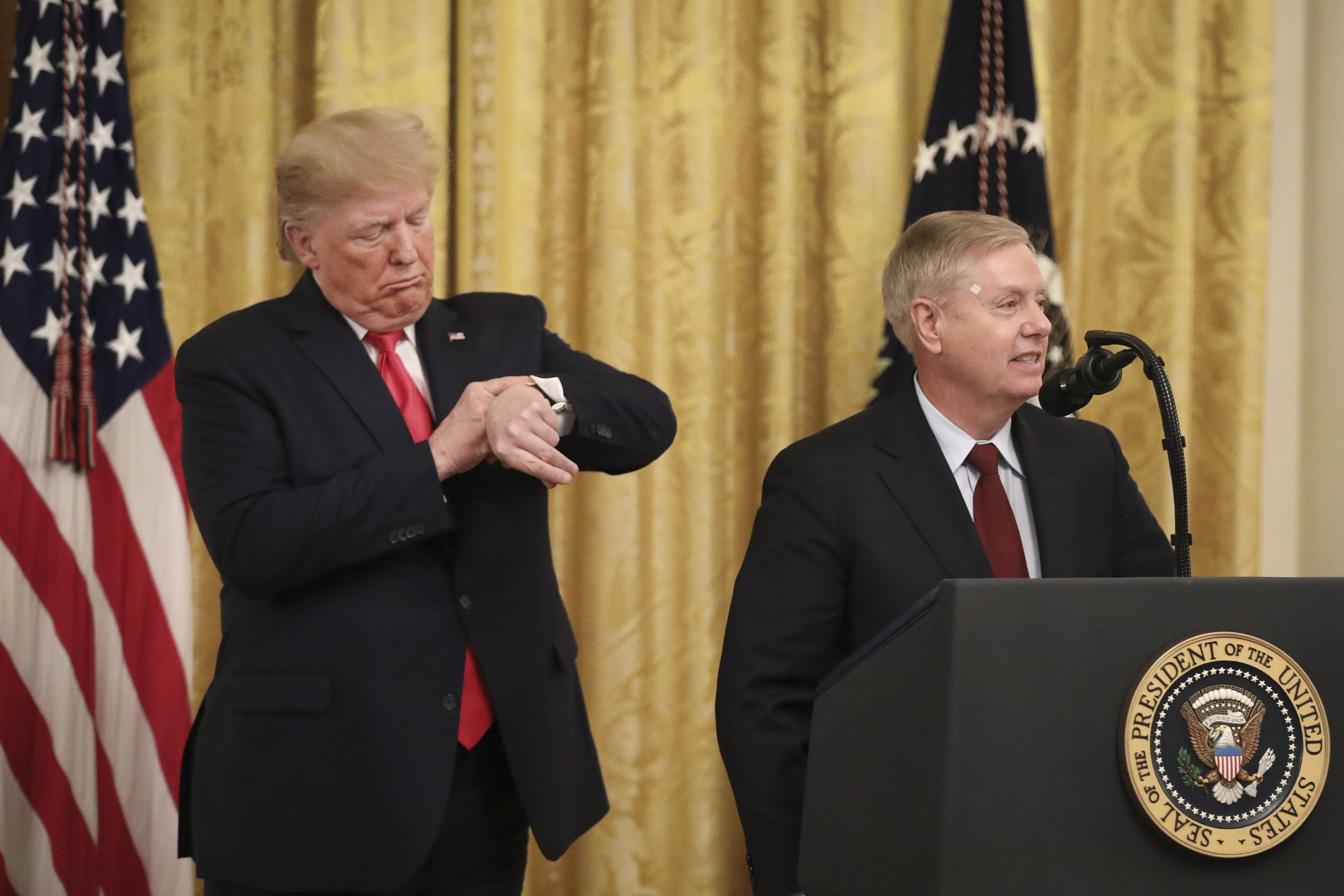 Two men in dark suits at a White House podium; one looks at his watch while the other speaks at a microphone in front of the Presidential seal.