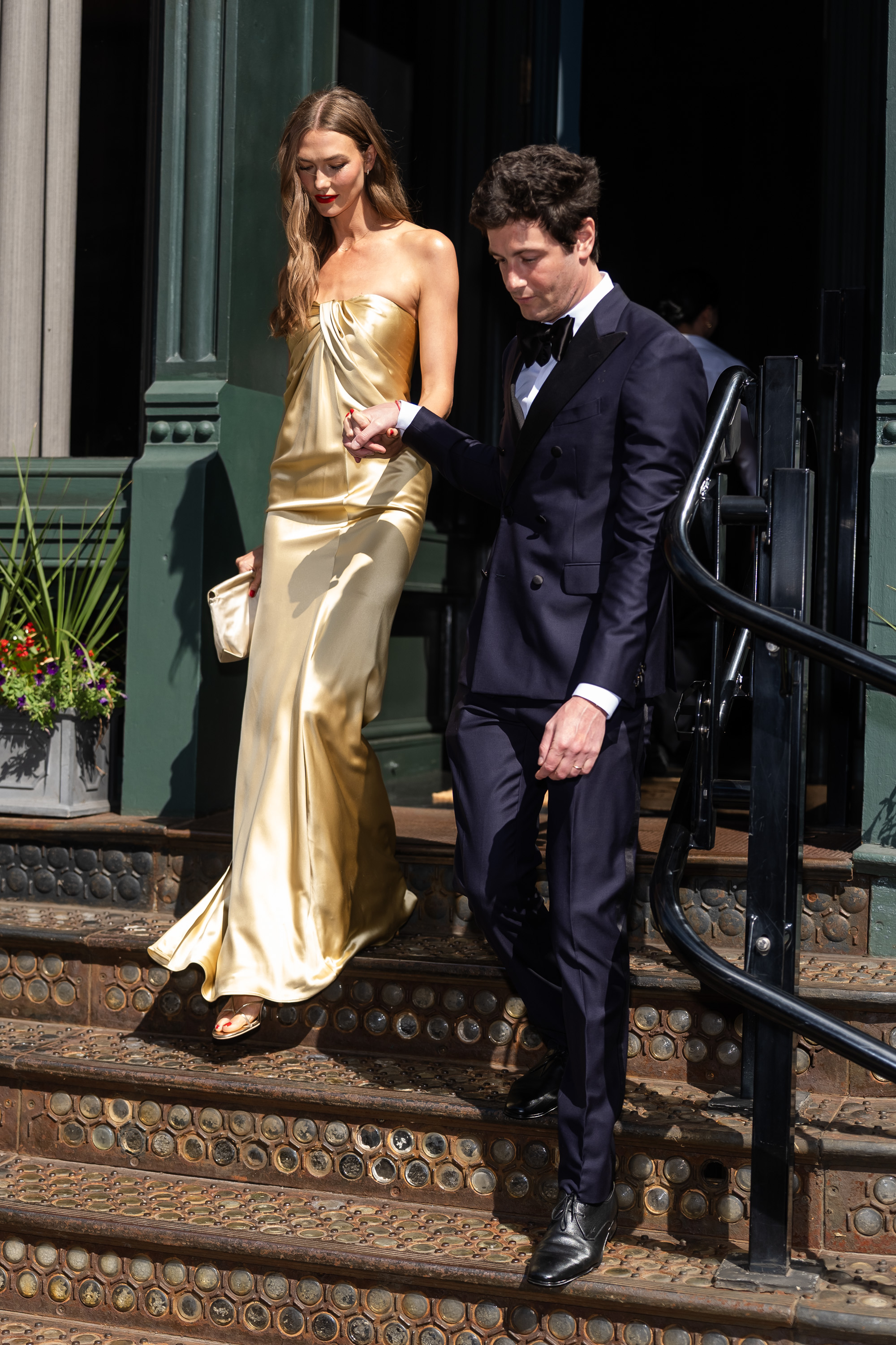 Karlie Kloss in a gold strapless satin gown and Joshua Kushner in a navy tuxedo walk down outdoor steps in SoHo, NYC.