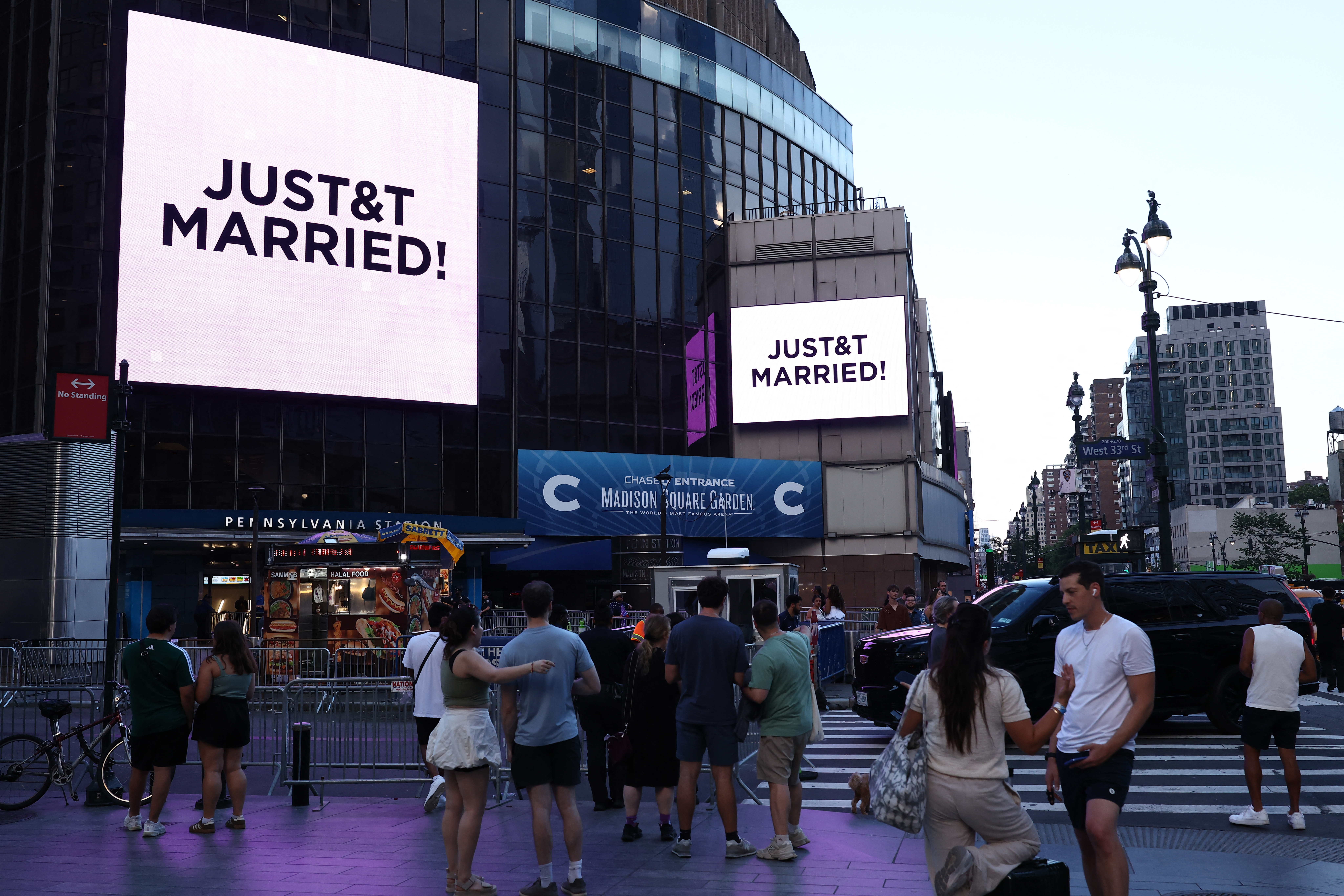 Crowd outside Madison Square Garden watching large screens displaying "JUST&T MARRIED!" celebrating Taylor Swift and Travis Kelce's wedding