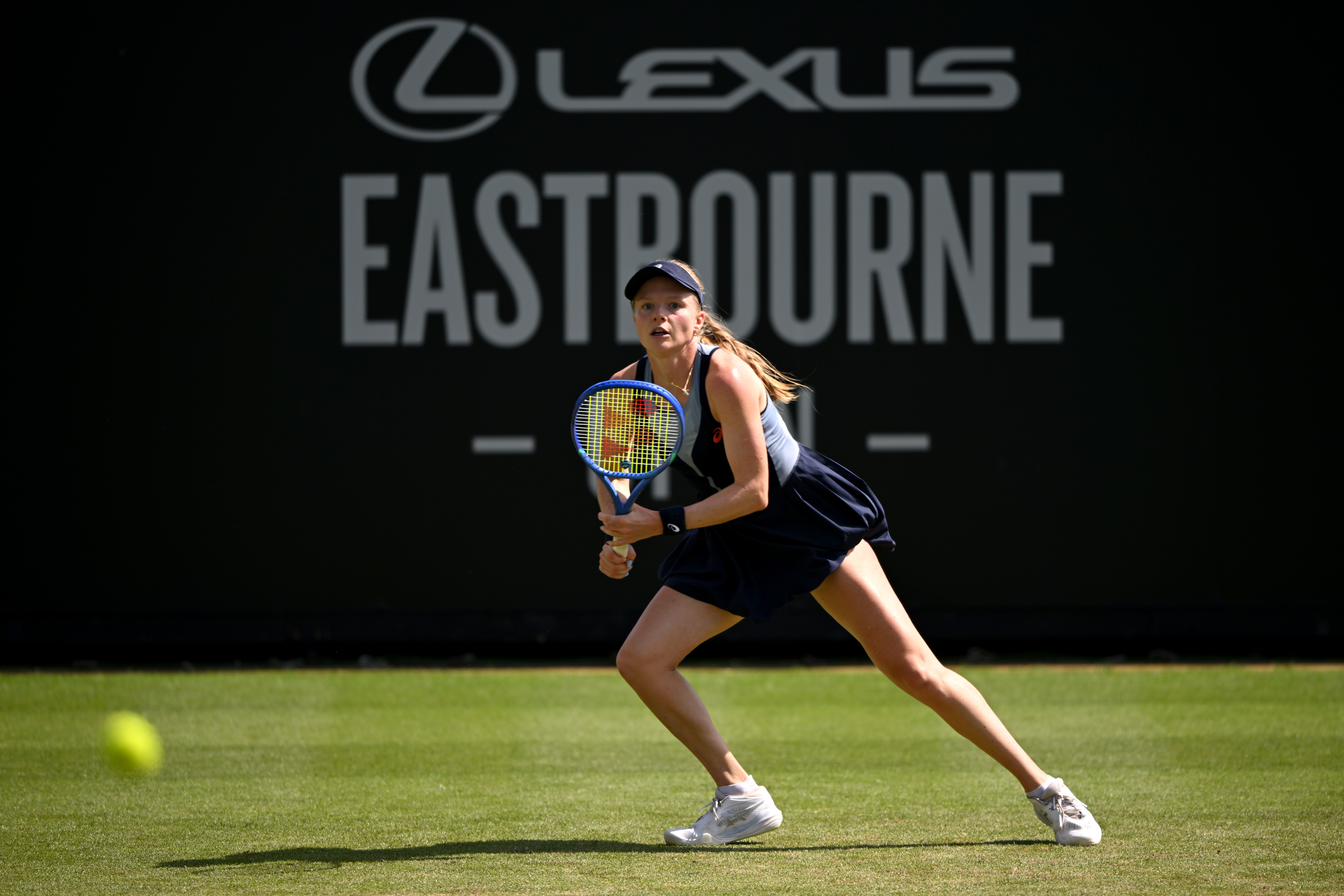 Female tennis player in navy outfit lunging forward on a grass court to return a shot, with a Lexus Eastbourne signboard in the background.