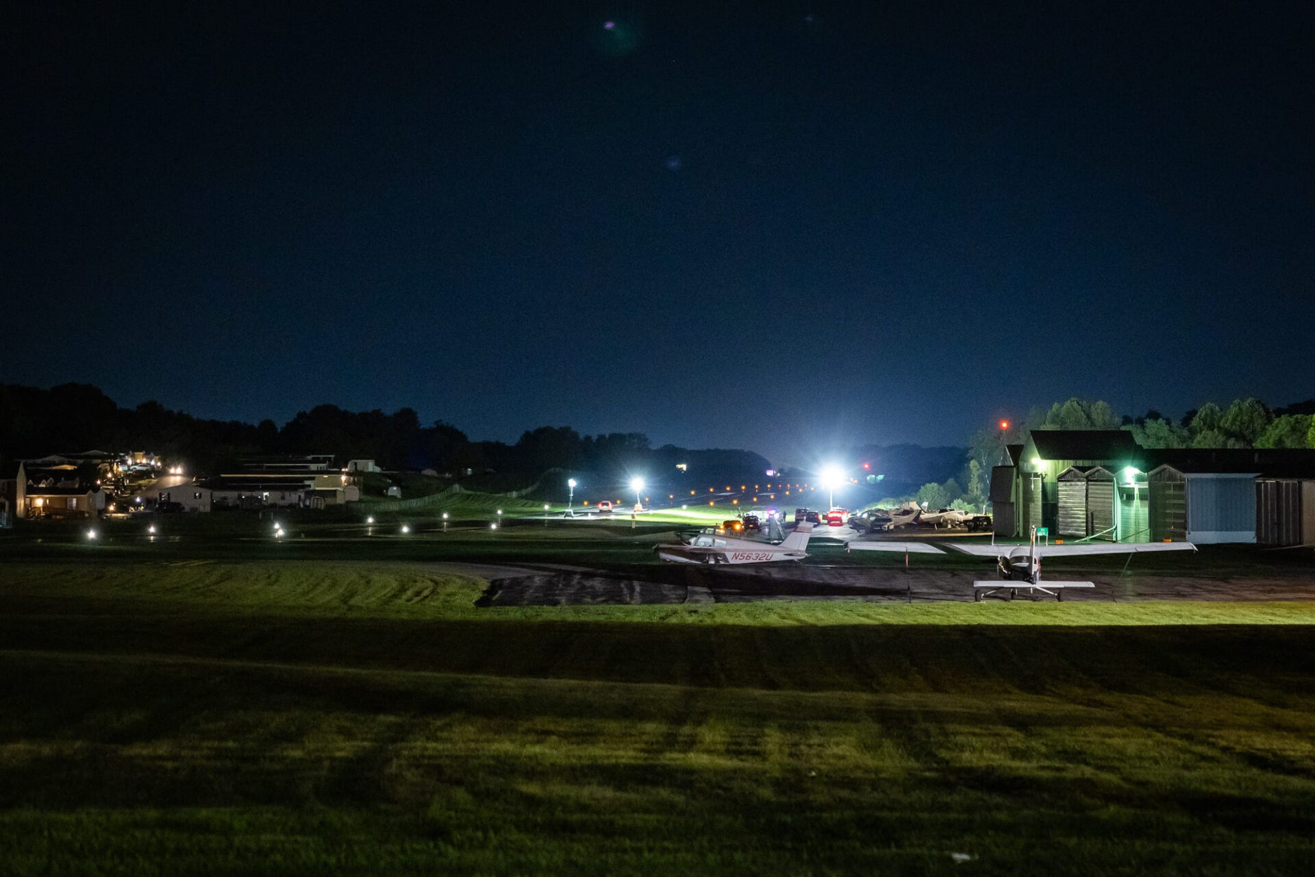 Wide nighttime shot of a small airport runway with emergency vehicle lights, parked small planes, hangars, and investigators visible in the distance.