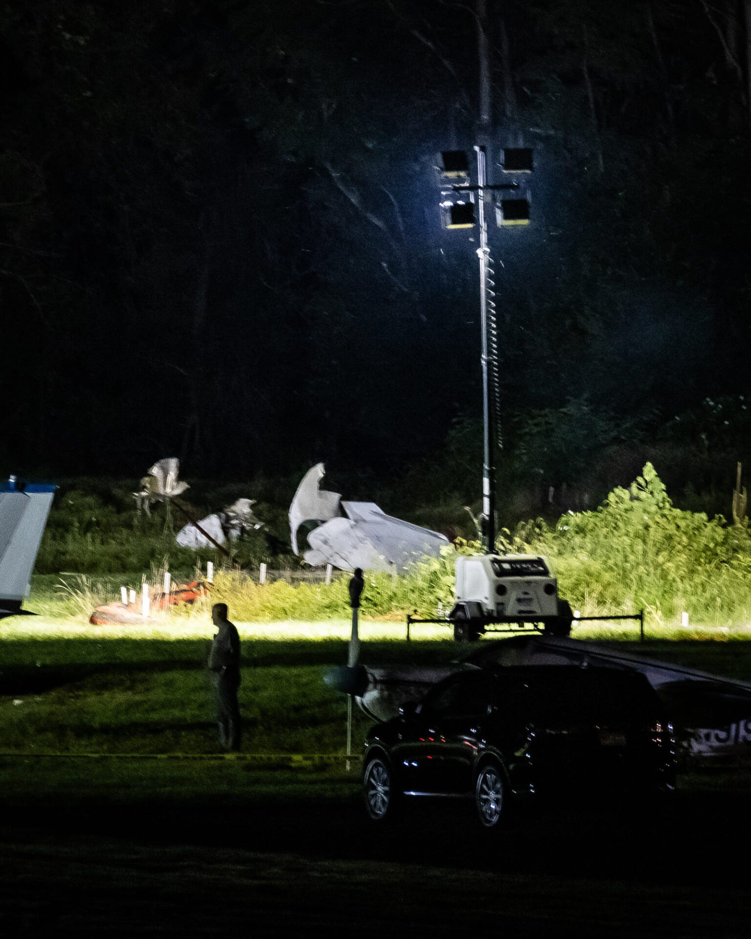 Crumpled white wreckage of a small plane strewn across a grassy area at night, illuminated by a portable light tower, with a person standing nearby.
