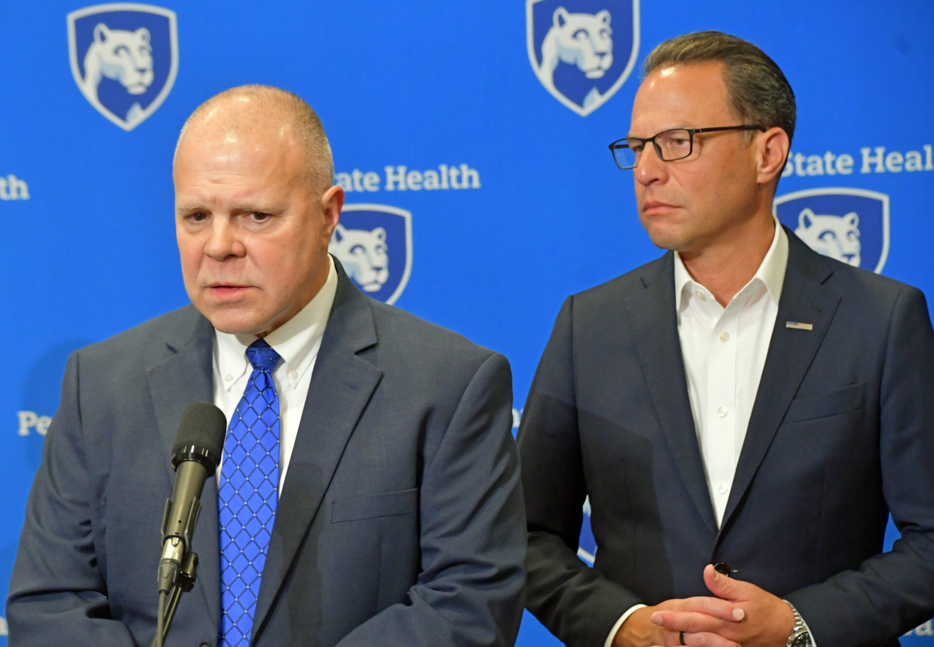 Lt. Col. George Bivens speaks at a microphone while Gov. Josh Shapiro stands behind him at a Penn State Health press conference backdrop.