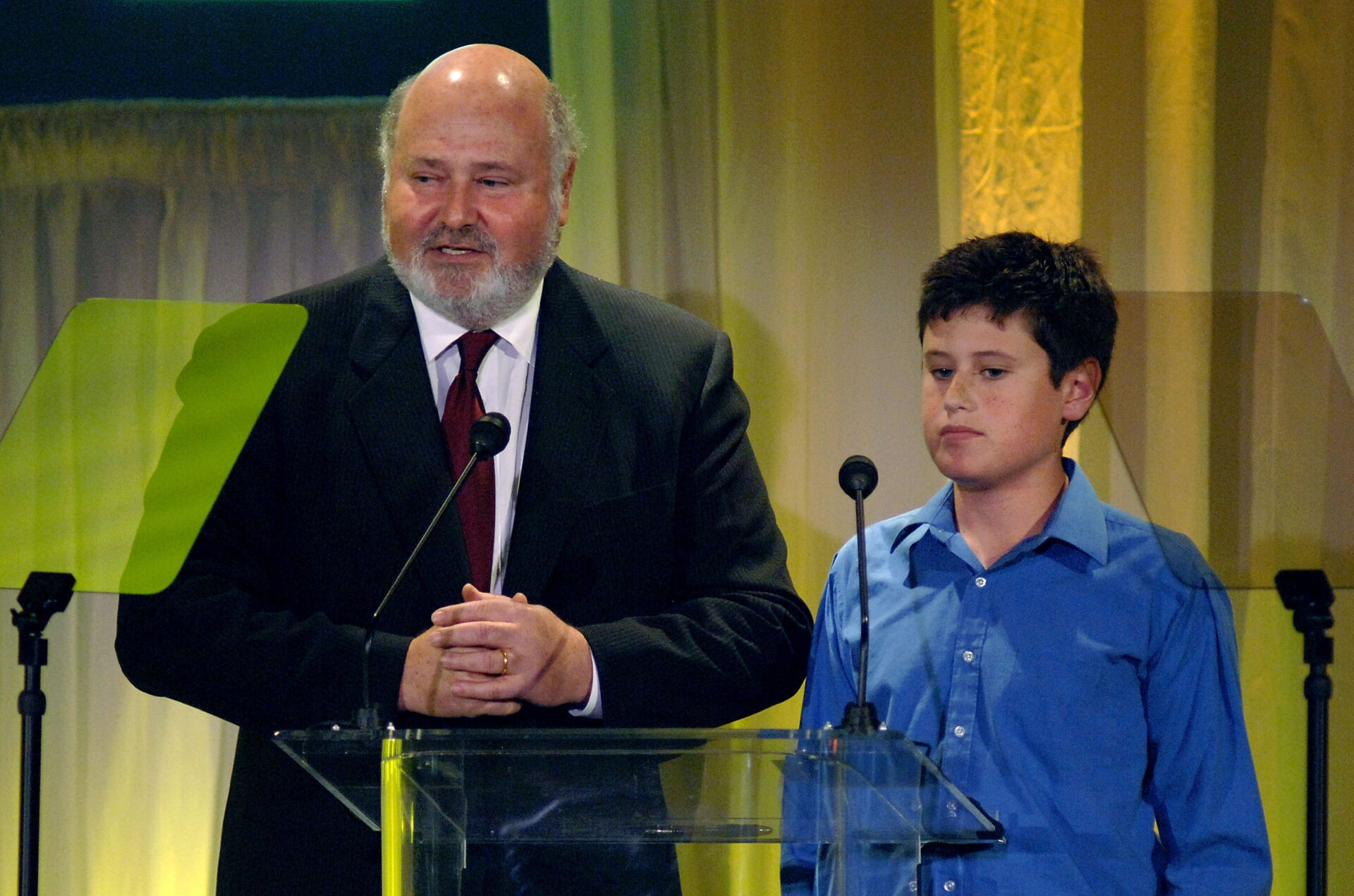 Bald older man in dark suit and red tie speaking at a glass podium alongside a young dark-haired boy in a blue shirt at an awards event.