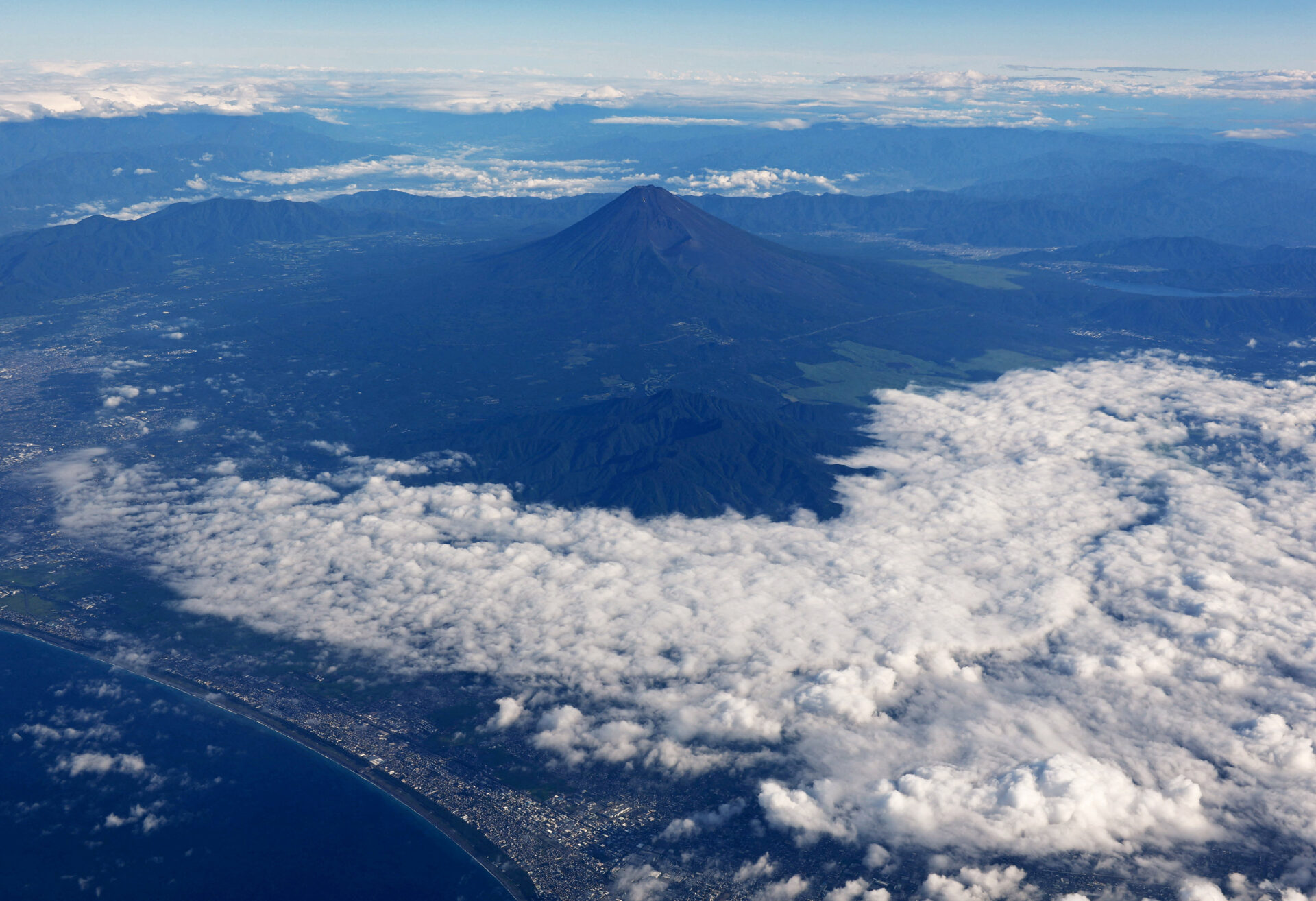 Aerial view of Mount Fuji's symmetrical volcanic cone rising above clouds, with coastline and surrounding landscape visible below.