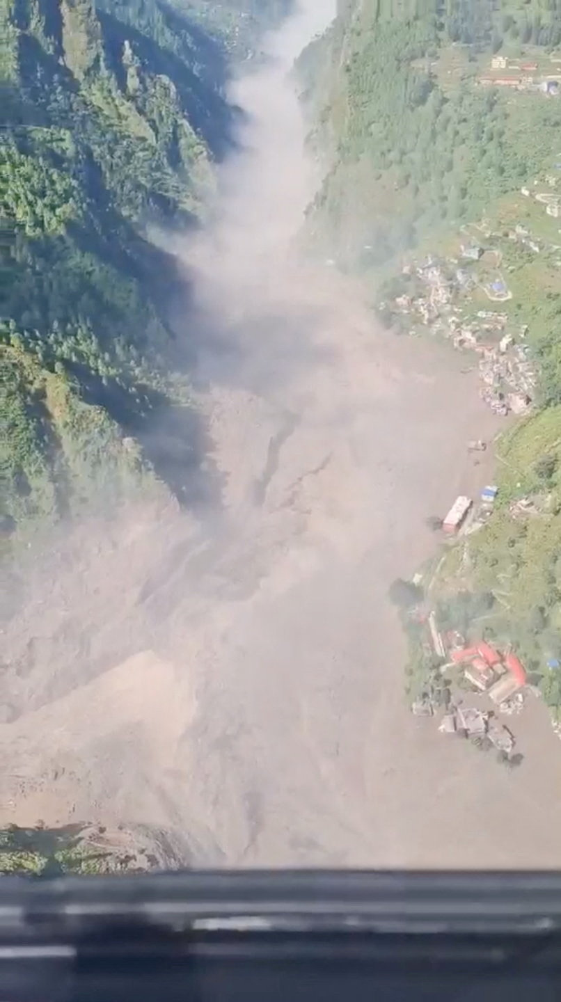 Wide aerial shot of a massive mudslide flowing between forested mountain slopes toward a riverside village in Rasuwa, Nepal.