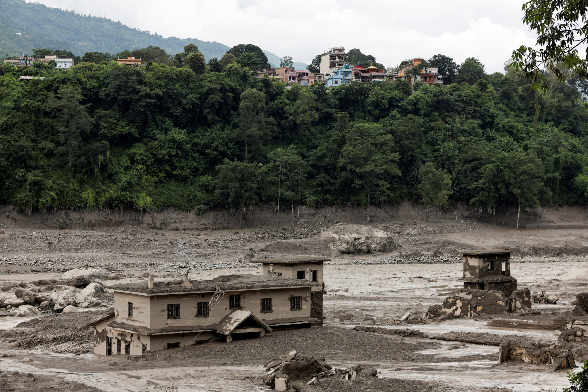 Mud-covered damaged buildings surrounded by flood debris in Trishuli, Nuwakot district, Nepal, with a hillside town visible in the background.