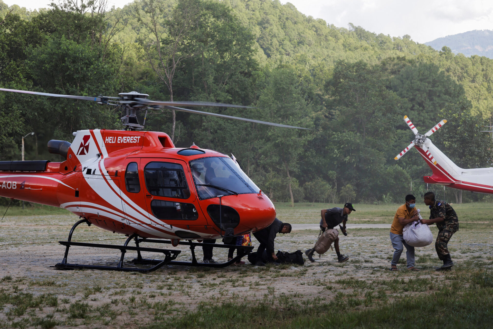 Workers and a soldier unloading bags from a red Heli Everest helicopter on a grass field at an army base in Nepal.