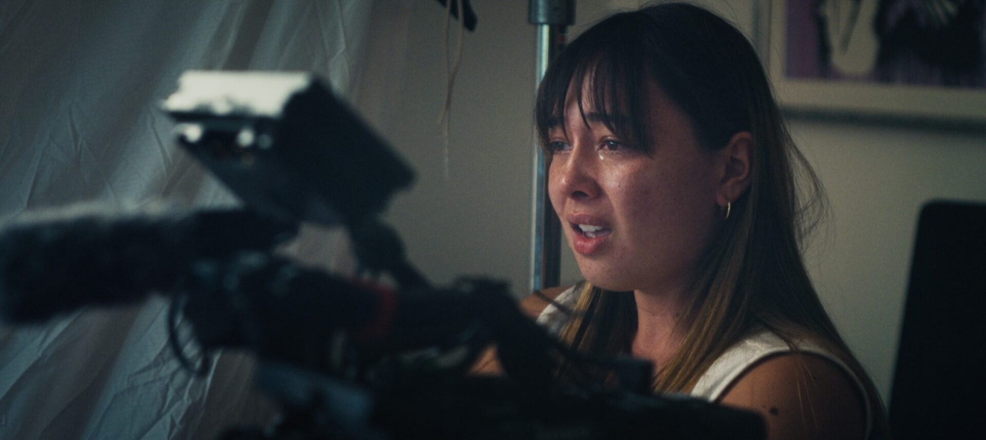 Young woman with dark bangs crying while seated next to a video camera on a rig, wearing a white tank top.