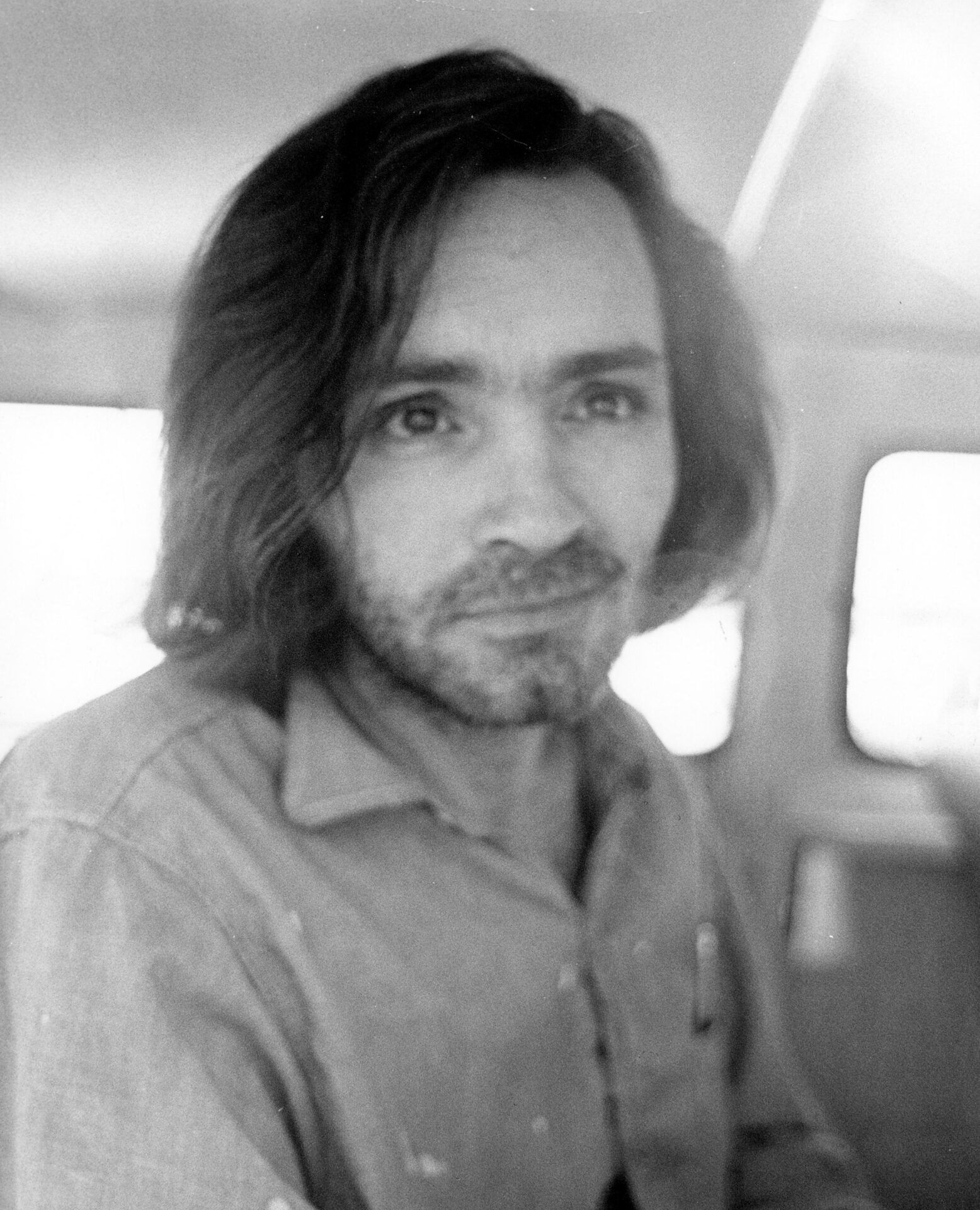 Black-and-white photo of Charles Manson with long hair and a beard, seated inside a police vehicle, wearing a collared shirt.