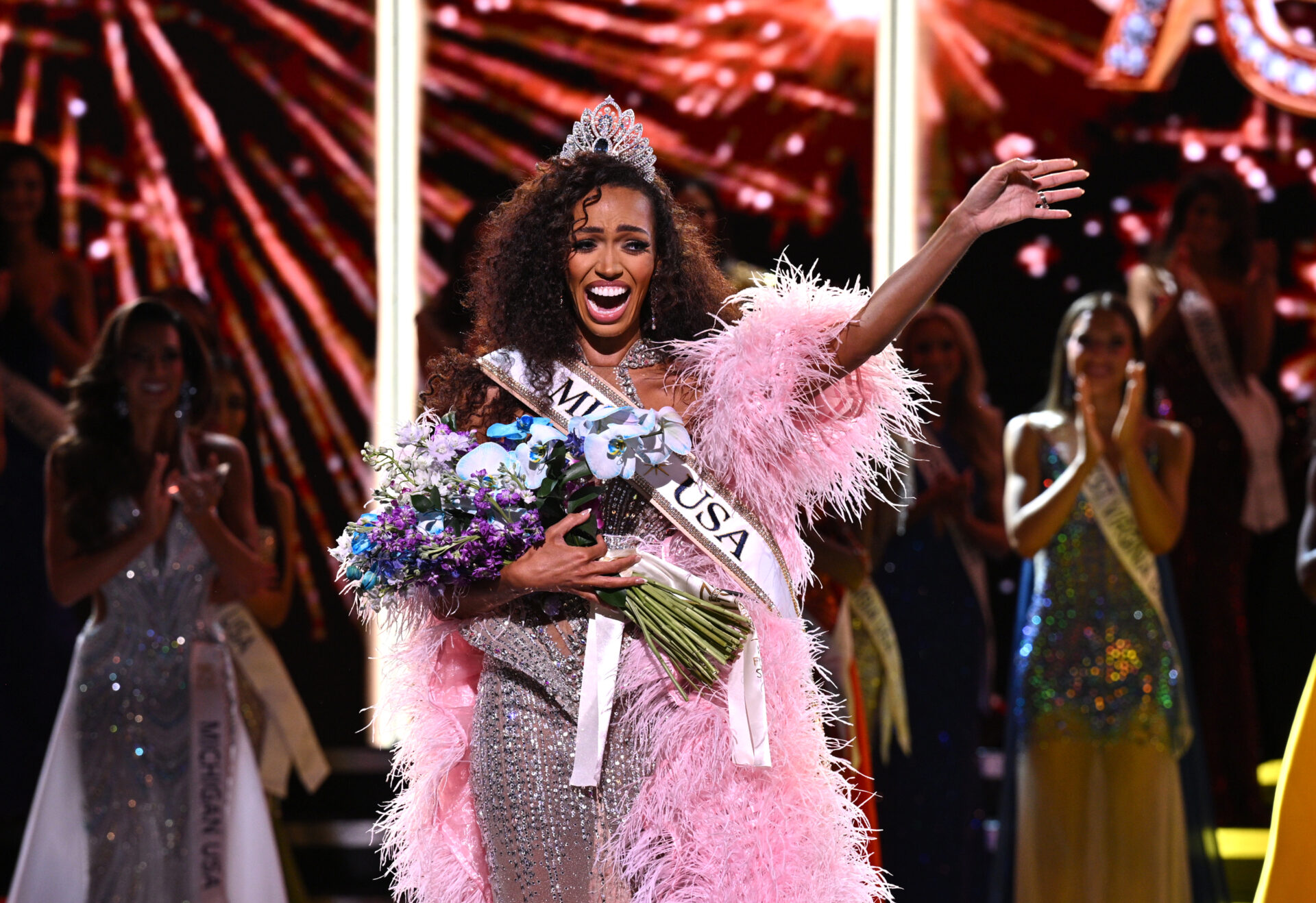 Miss USA 2026 Justus Kelley in a pink feathered gown, wearing a crown and sash, holding a bouquet and waving on stage.