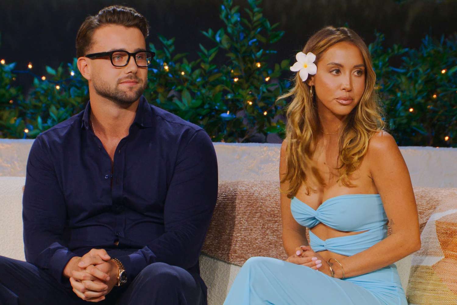 A man in a navy shirt and glasses and a woman in a light blue strapless outfit with a white flower in her hair sit together on a couch outdoors.