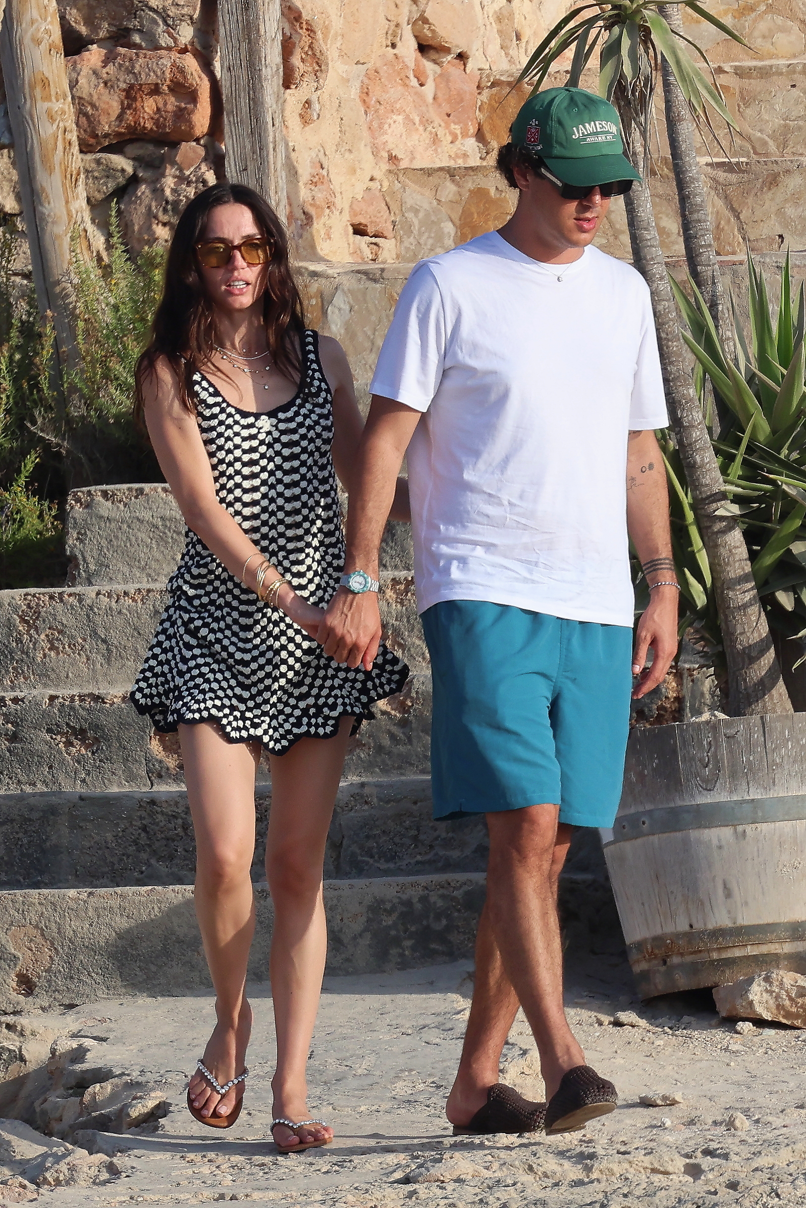 A woman in a black and white mini dress and sunglasses holds hands with a man in teal shorts and a green Jameson cap while walking on a beach.