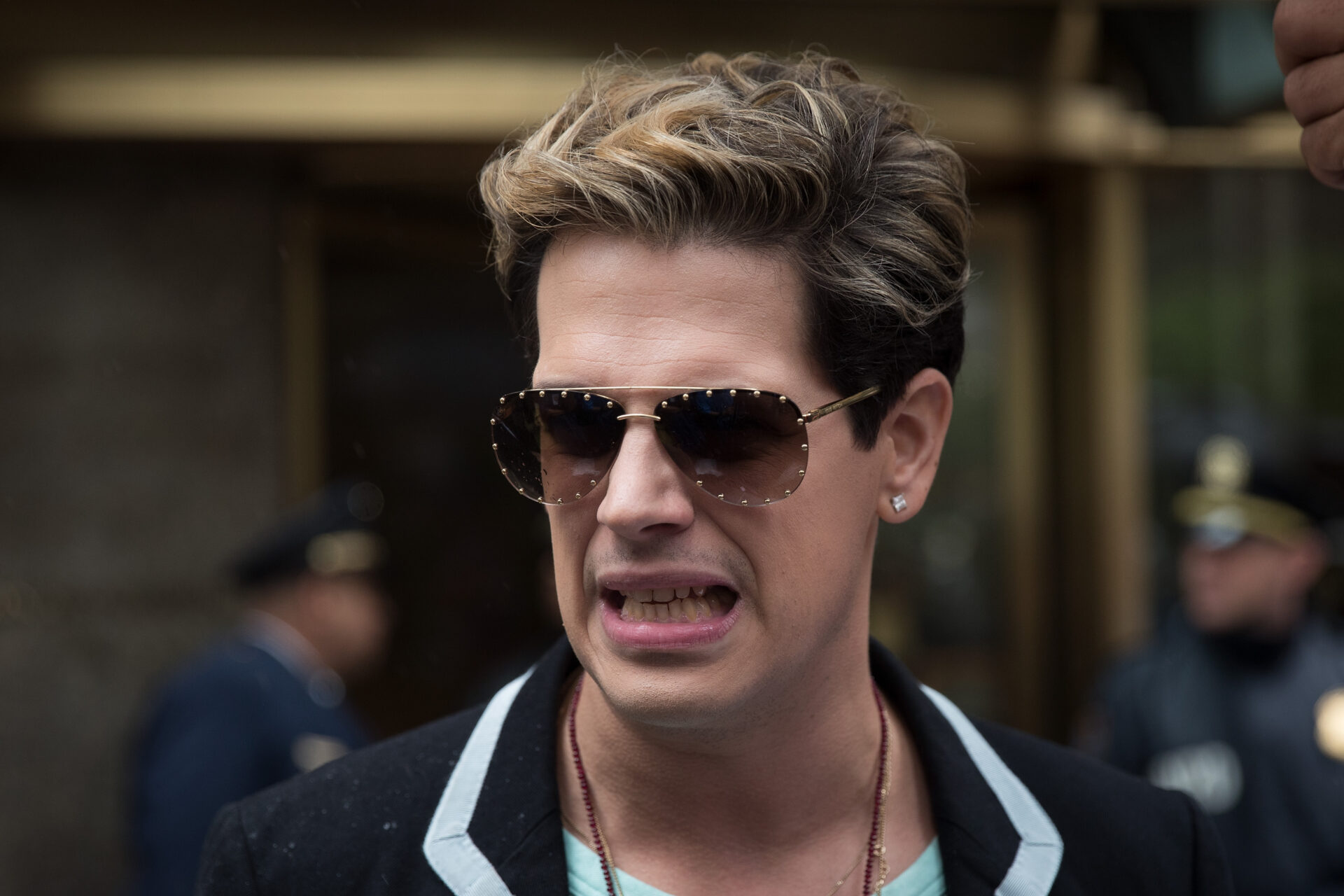 Man with blond-streaked hair and aviator sunglasses speaking outside a building, mouth open, with police officers blurred in the background.