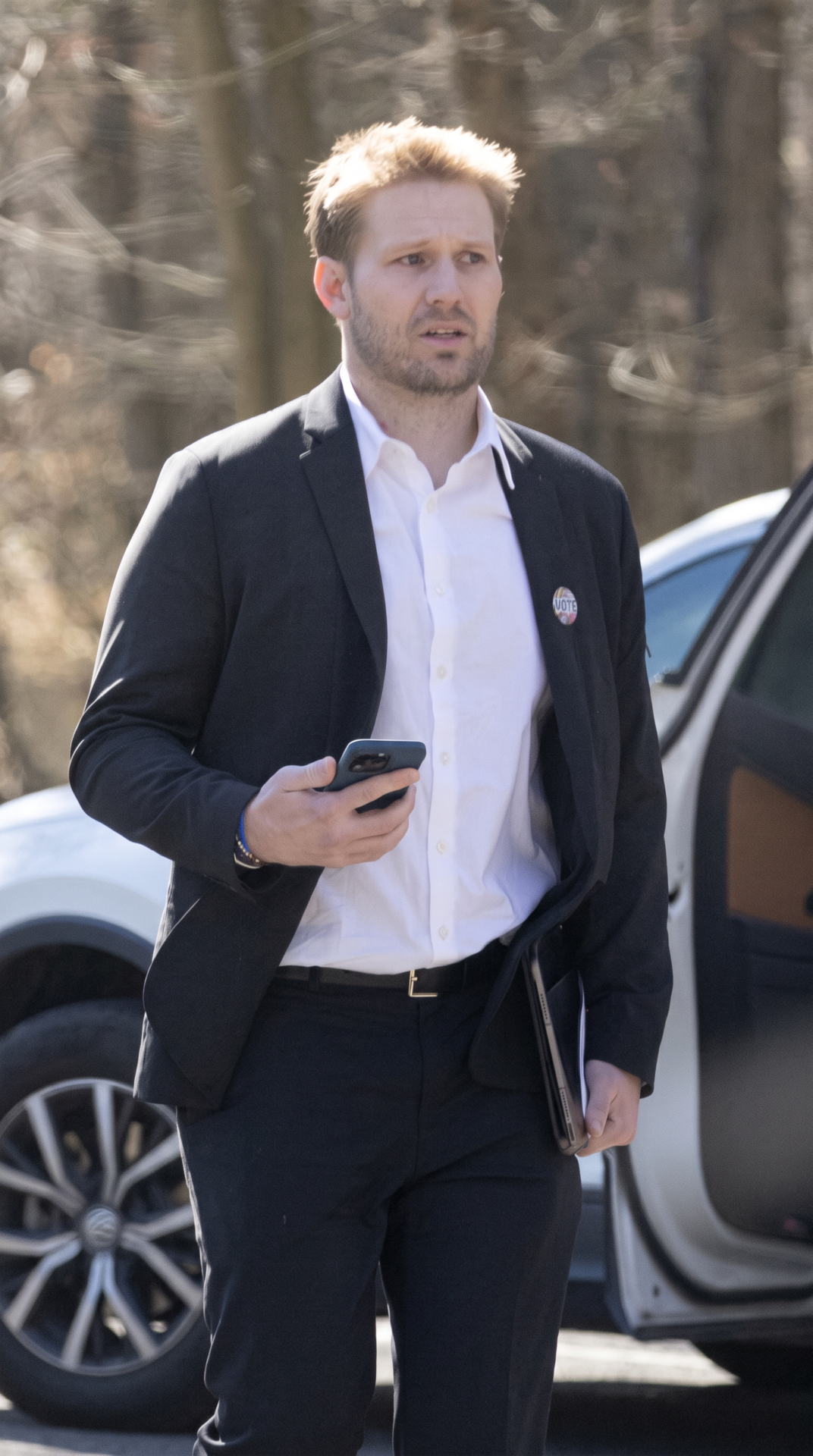 Man in a dark blazer and white shirt holding a smartphone, walking near a white SUV with bare trees in the background.