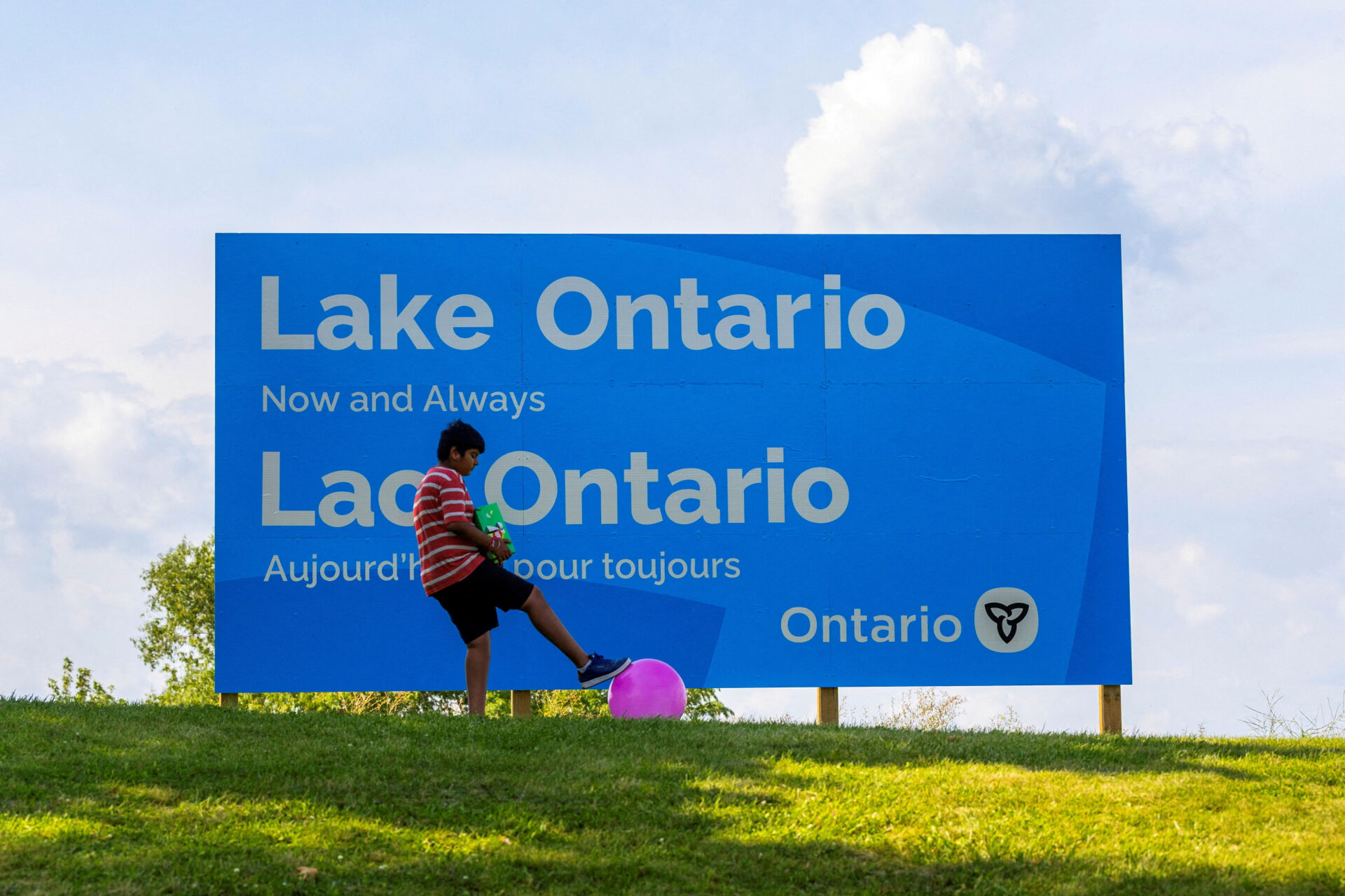 A young boy in a red striped shirt kicks a pink ball in front of a large blue billboard reading "Lake Ontario, Now and Always / Lac Ontario, Aujourd'hui pour toujours" with the Ontario government logo.