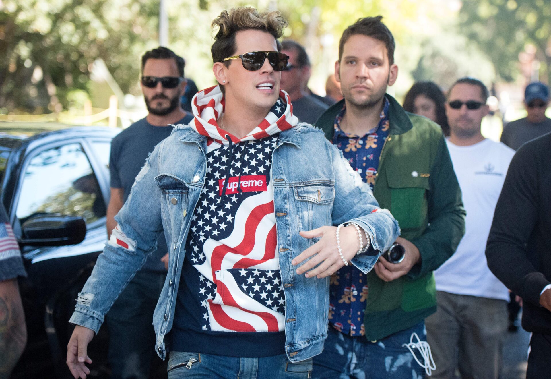 Man in sunglasses, American flag Supreme hoodie, and distressed denim jacket walks through a crowd outdoors near a parked car.