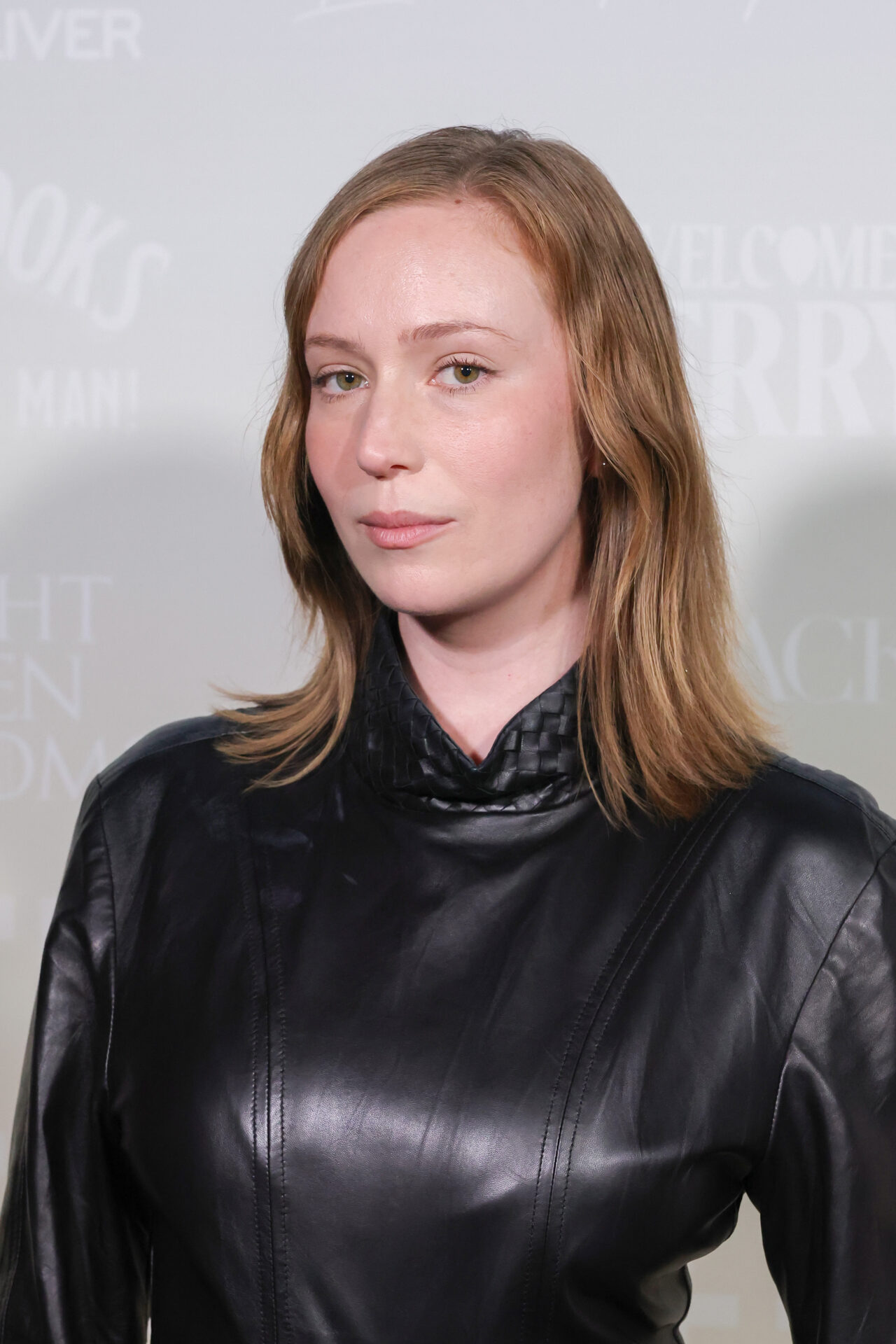 Woman with medium-length auburn hair in a black leather turtleneck top posing against a light event backdrop.