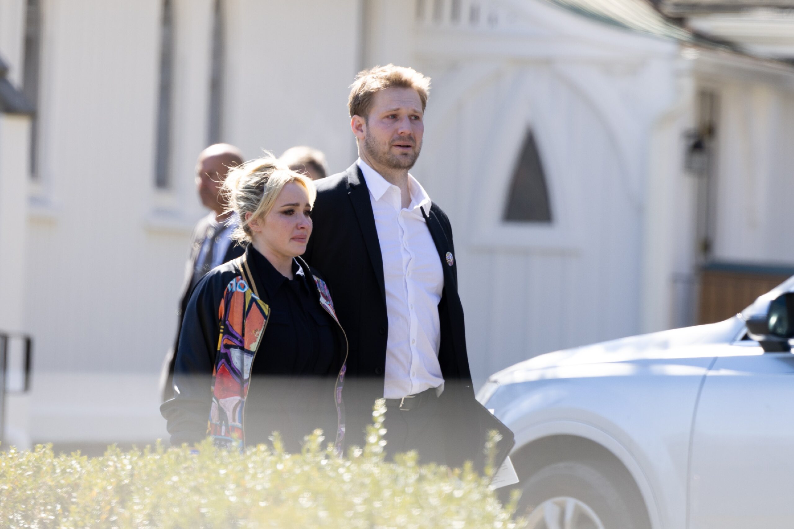 A visibly emotional blonde woman in a colorful jacket walks alongside a man in a white shirt and dark blazer near a white building.
