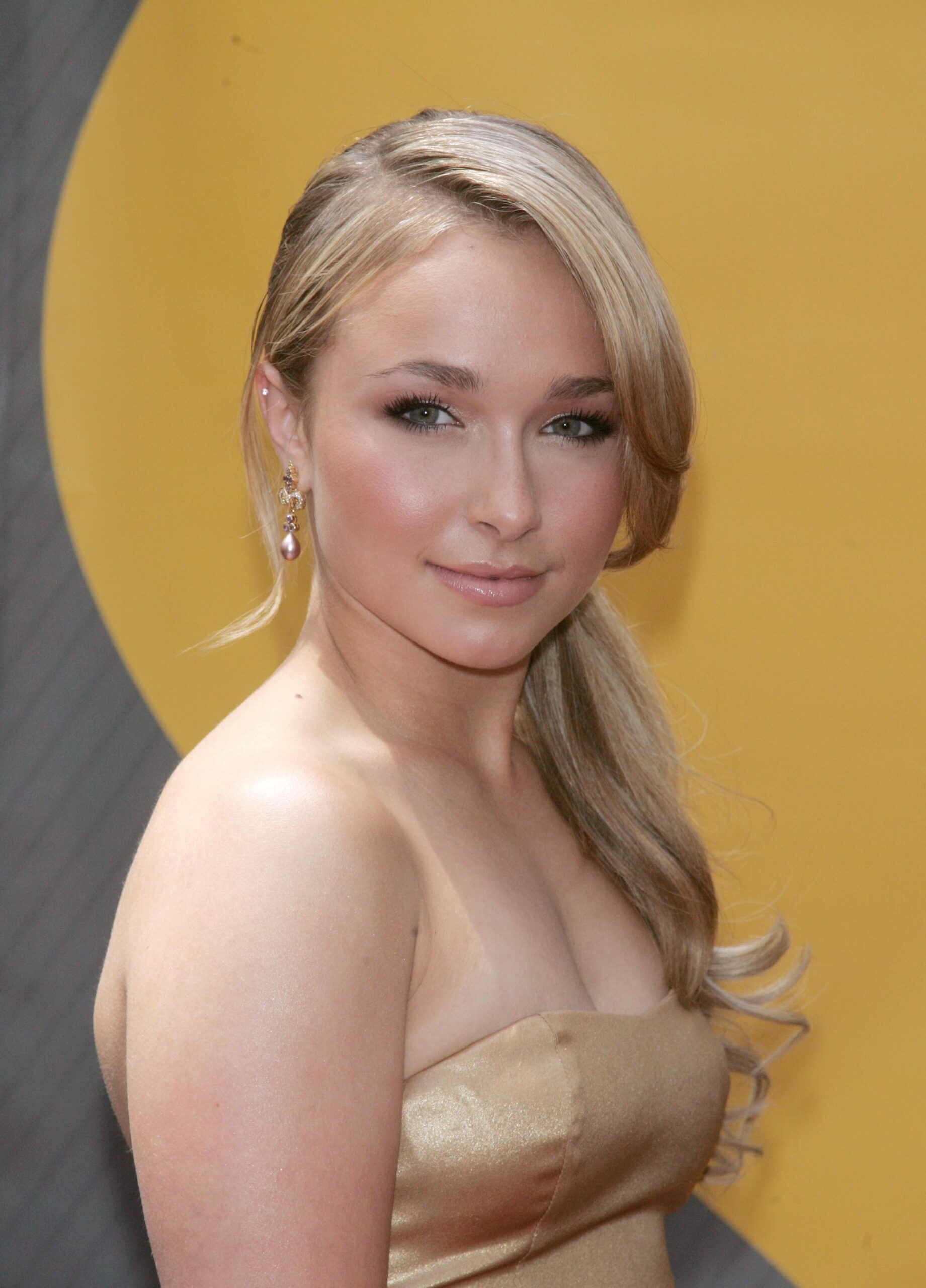 Young blonde woman in a gold strapless dress with sleek side-swept hair posing against a yellow circular backdrop.